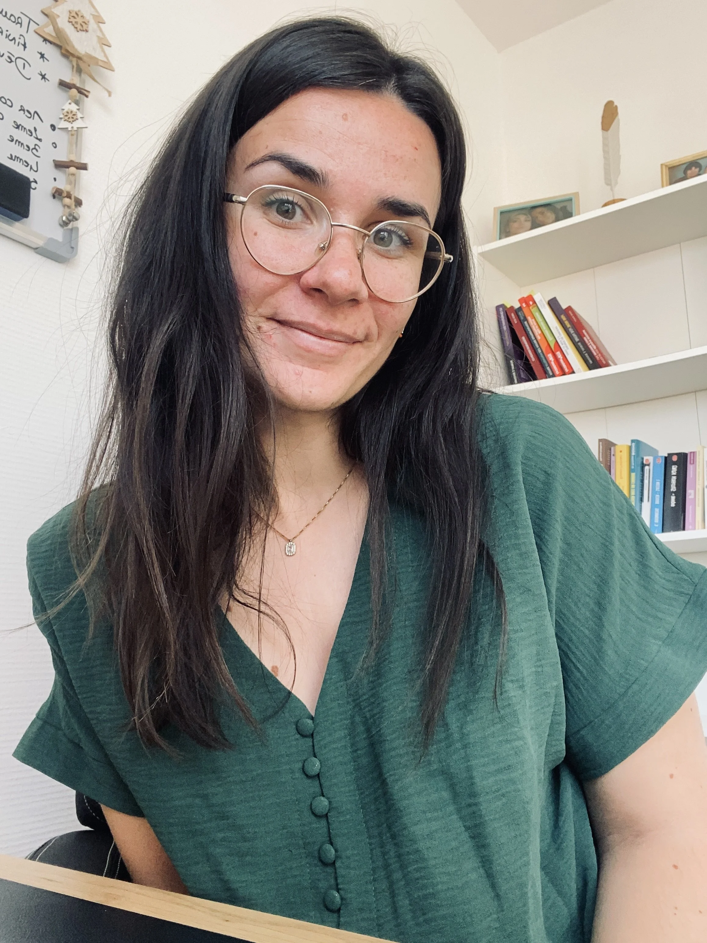 Une femme avec des lunettes rondes, des cheveux longs et noirs, portant une chemise verte, sourit à la caméra dans une pièce avec des étagères de livres et des décorations en arrière-plan.