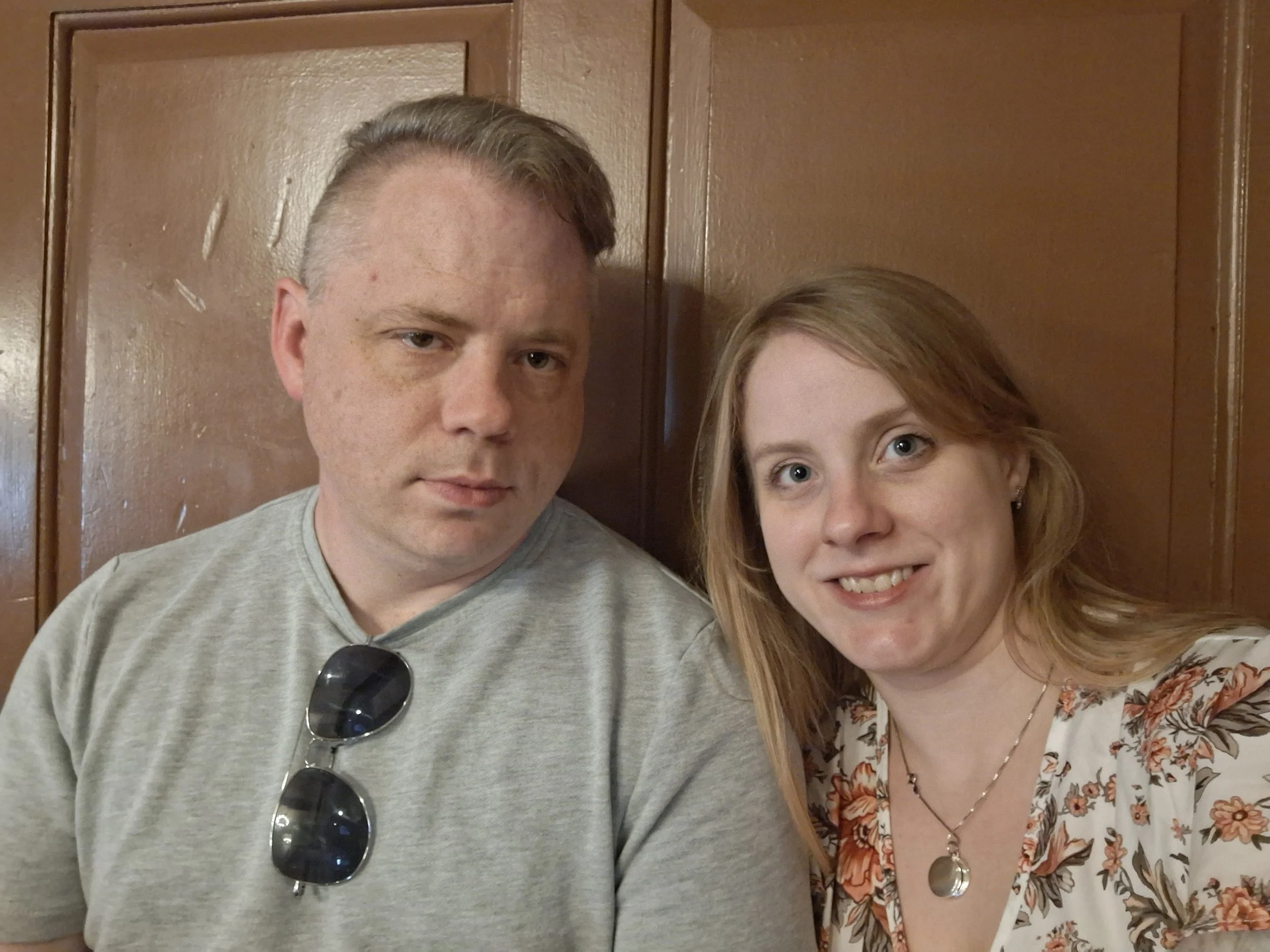 A man and woman sit close together, in front of a brown wooden wall. The man has short light brown hair and is wearing a gray t-shirt with sunglasses hanging from the collar. The woman has shoulder-length reddish hair and is smiling, wearing a floral blouse with a necklace.