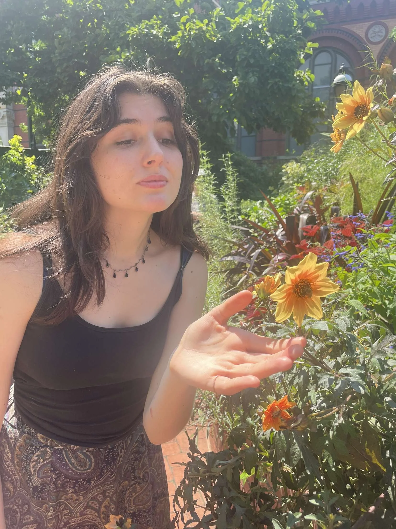 A young woman with long brown hair and light skin stands outdoors in a garden on a sunny day, near colorful flowers including yellow and orange blooms, with a red brick building in the background.