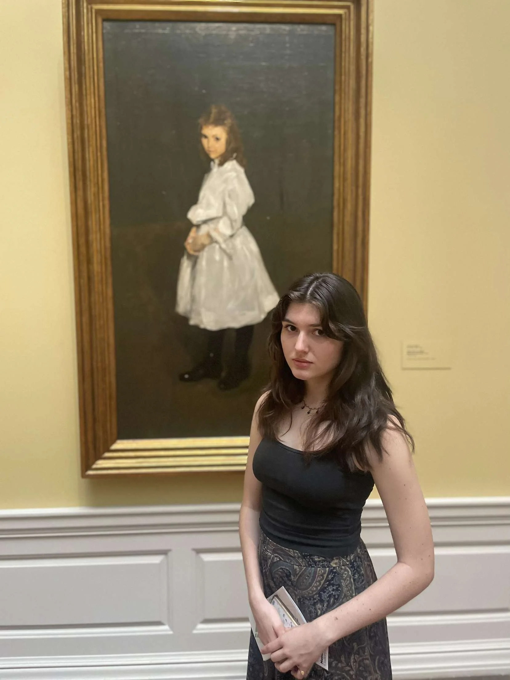 A young woman with wavy brown hair standing in front of a painted portrait of a girl in a white dress in an art gallery.