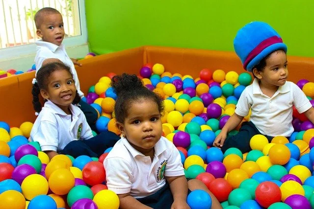 Four young children playing in a colorful ball pit with green walls and window.