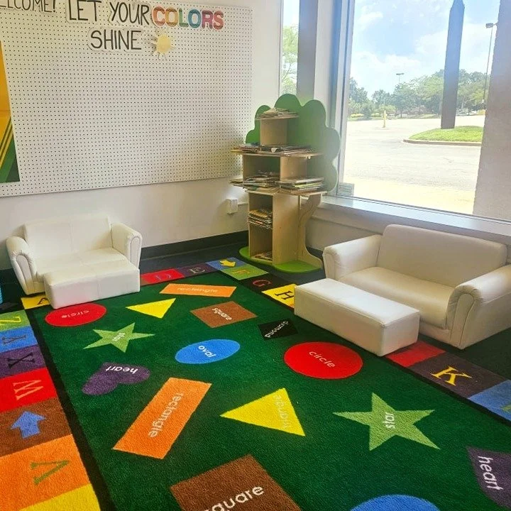 Colorful playroom corner with white sofas, a green bookshelf shaped like a tree, a large window, and a vibrant carpet with letters, shapes, and words.