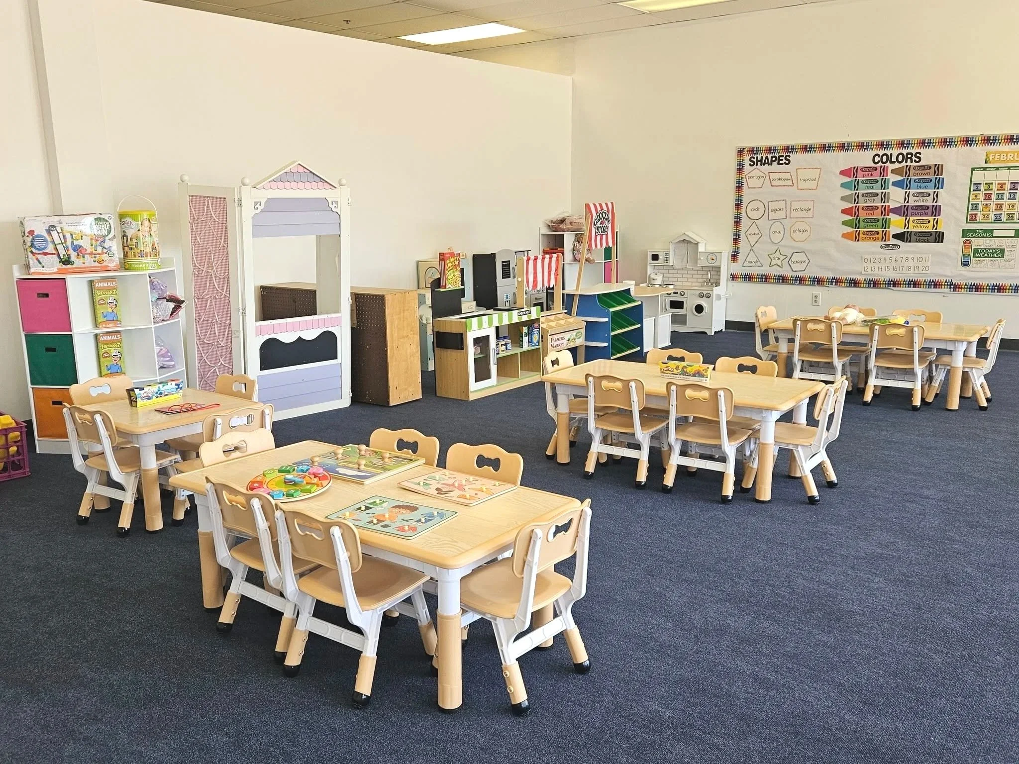 A preschool classroom with small wooden tables and chairs, educational toys, a pretend playhouse, and a colorful shape and color chart on the wall.