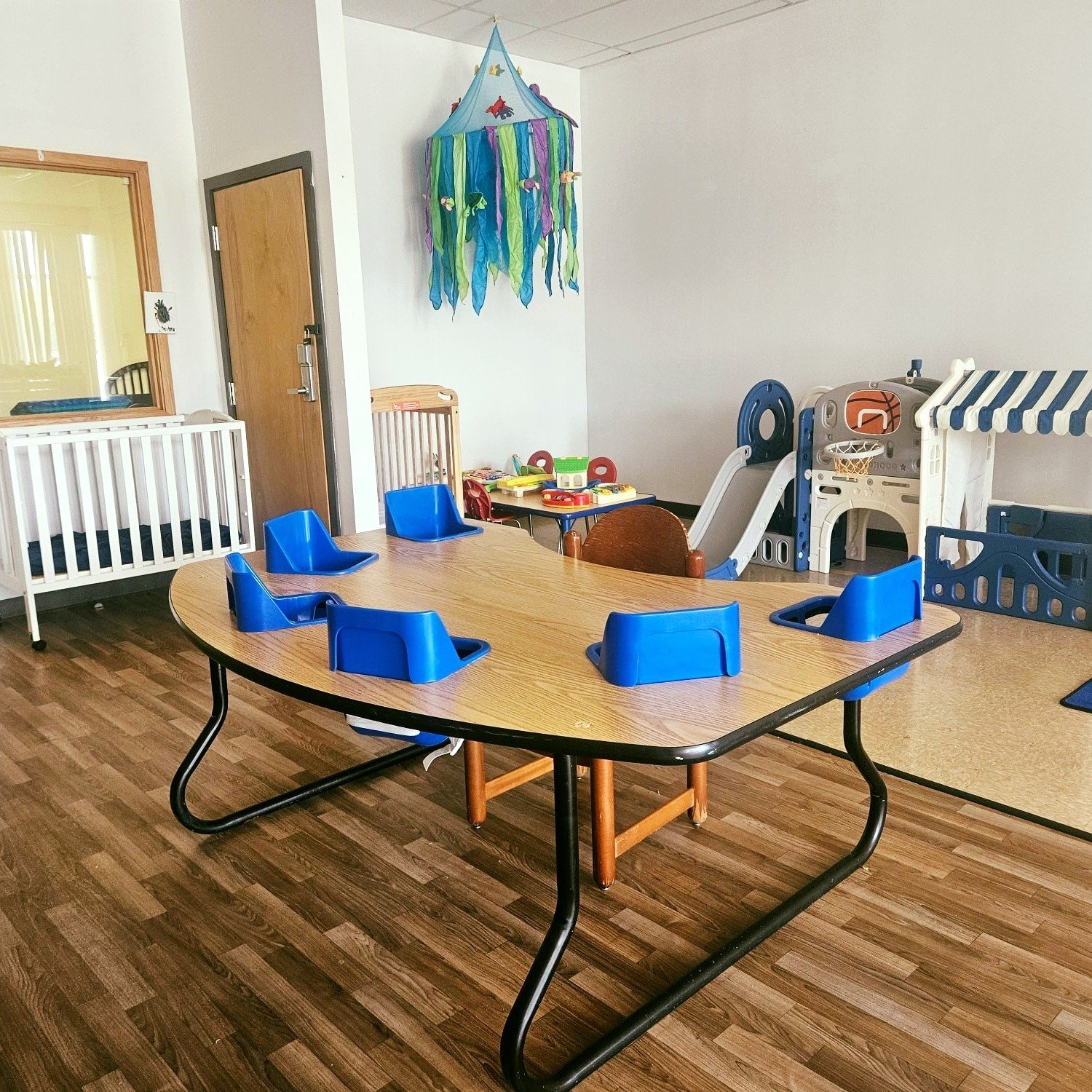 A classroom with a wooden table with blue high chairs, a small play area with miniature sports equipment, and a colorful hanging wall decoration resembling a jellyfish. There is a white crib and a playhouse with a slide in the background.