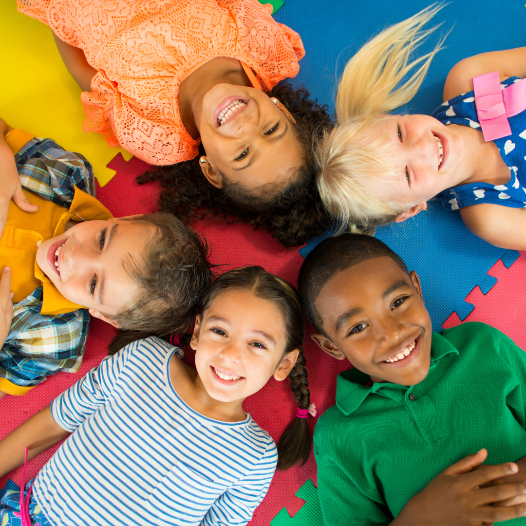 Six children lying in a circle on colorful foam mats, smiling and looking up at the camera.