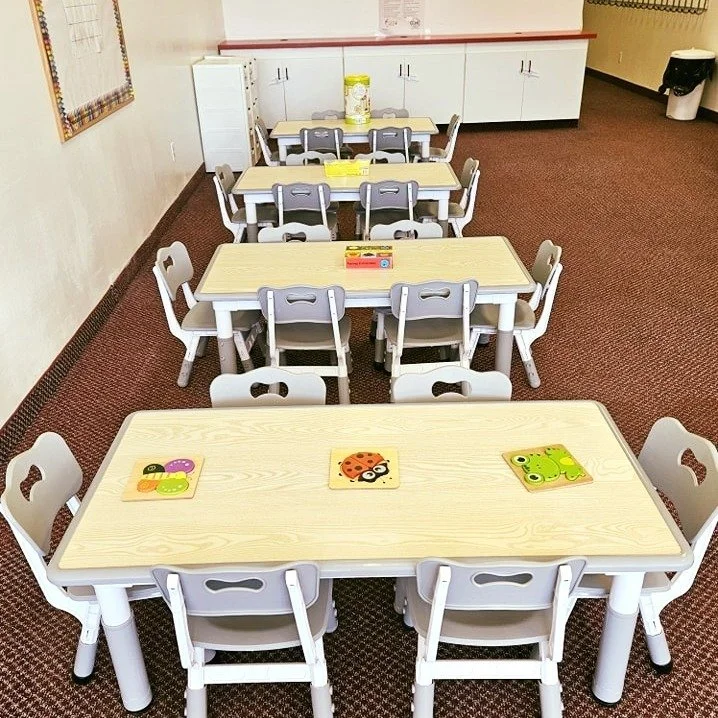 Three empty rectangular tables with children’s chairs are arranged in a classroom or daycare setting. The tables have picture cards, including a ladybug, ladybug with flowers, and frogs, placed on top. In the background, there are cabinets, a container on the cabinet, and a trash bin in the corner.