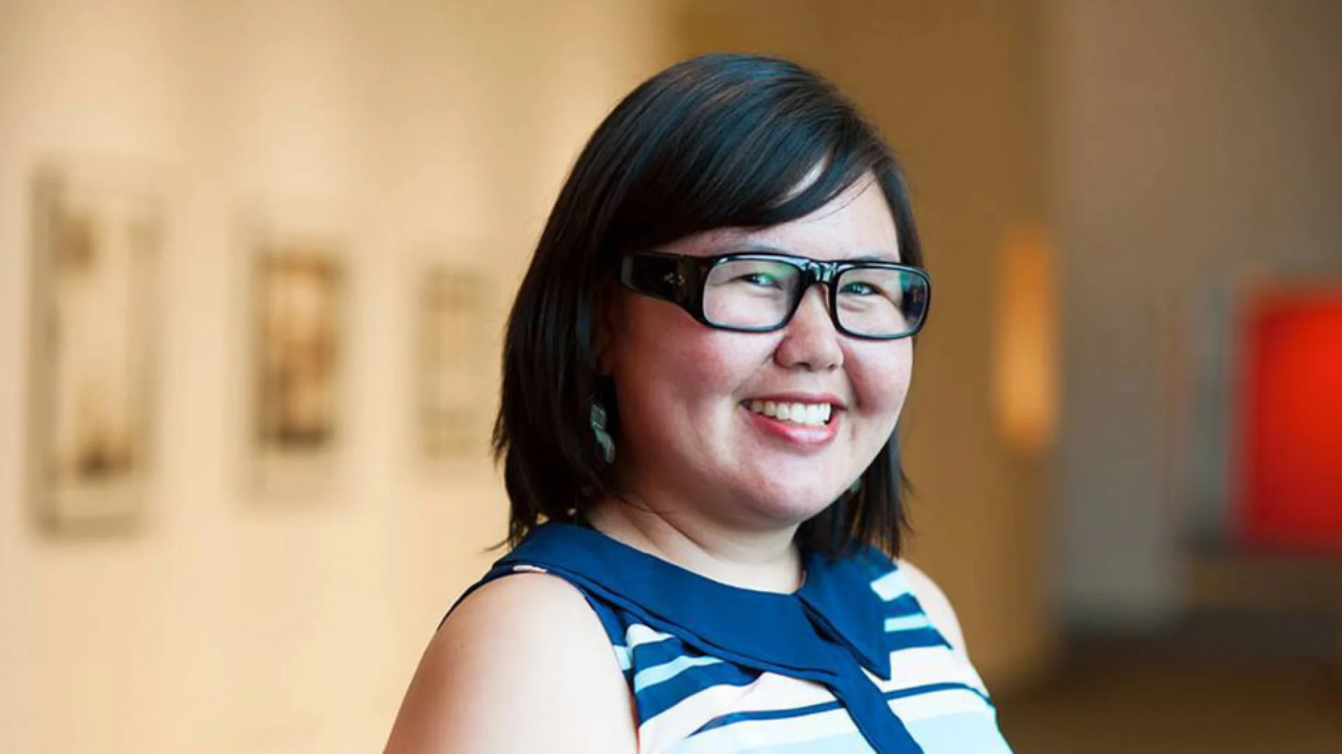A woman with shoulder-length black hair, wearing black glasses, smiling, in an indoor setting with blurred artwork in the background.