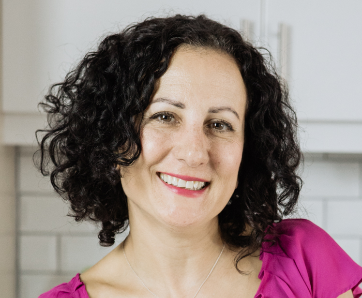 A woman with shoulder-length curly dark hair smiling, wearing a pink top in a kitchen setting.