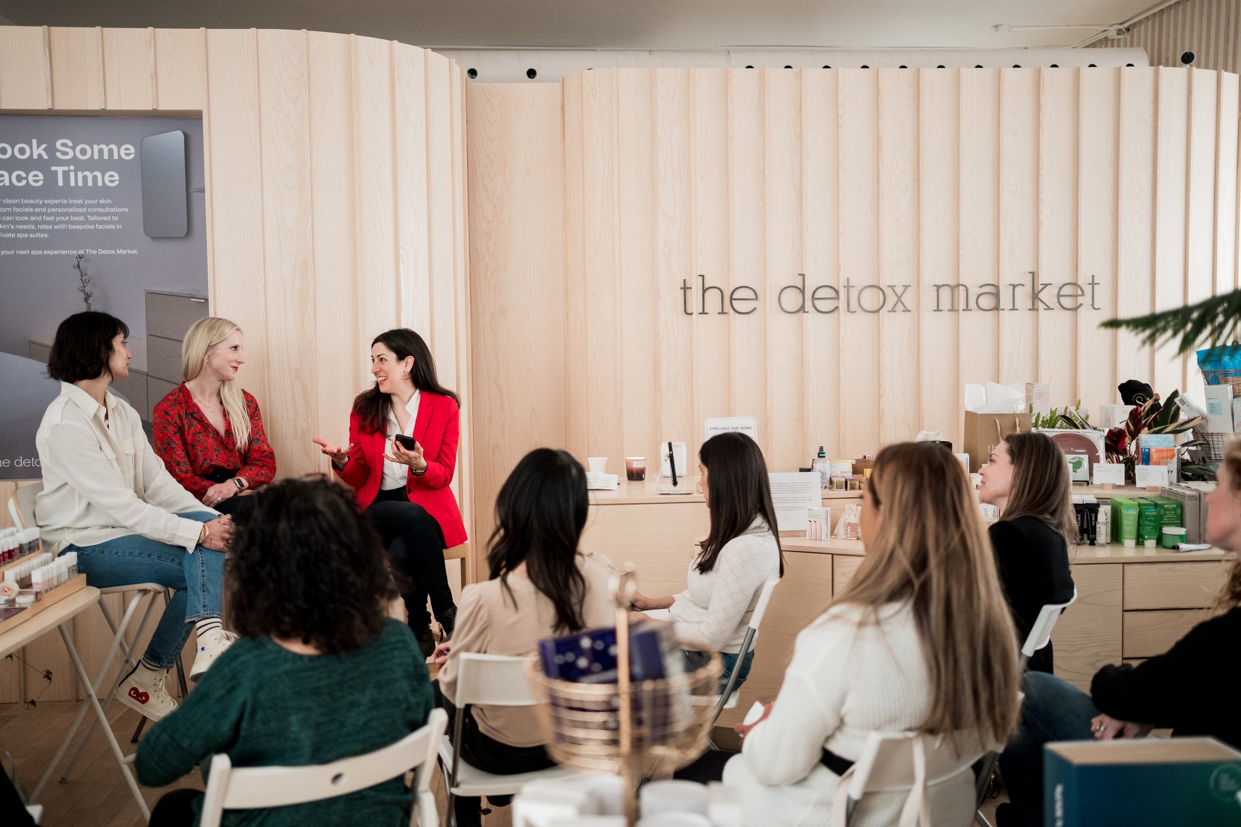 Group of women participating in a discussion at the Detox Market, with three women seated on a small stage and a group of women seated in front of them, inside a wellness store with light wood decor.
