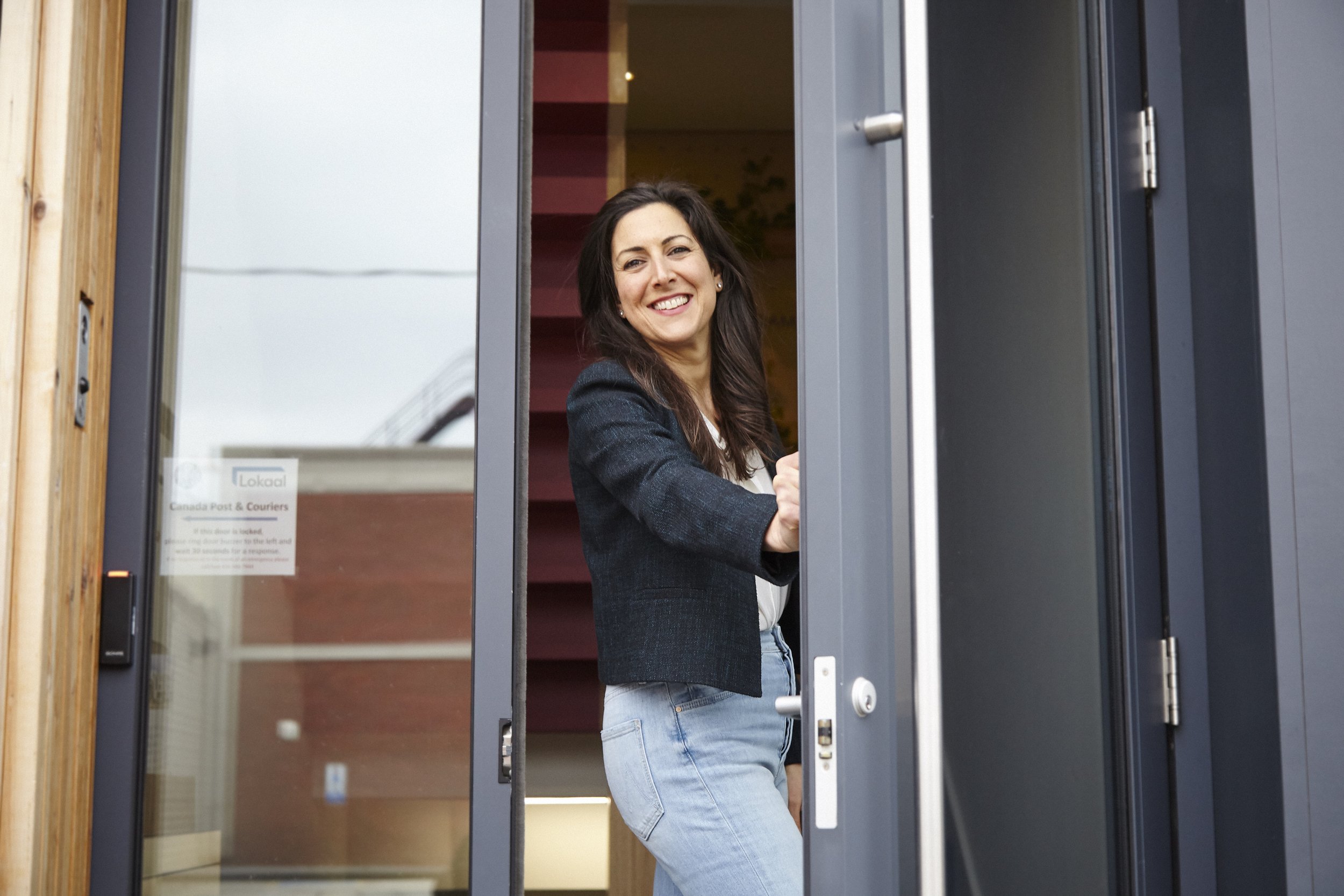 A woman with long dark hair wearing a black blazer and jeans, smiling and standing in a doorway, looking out.