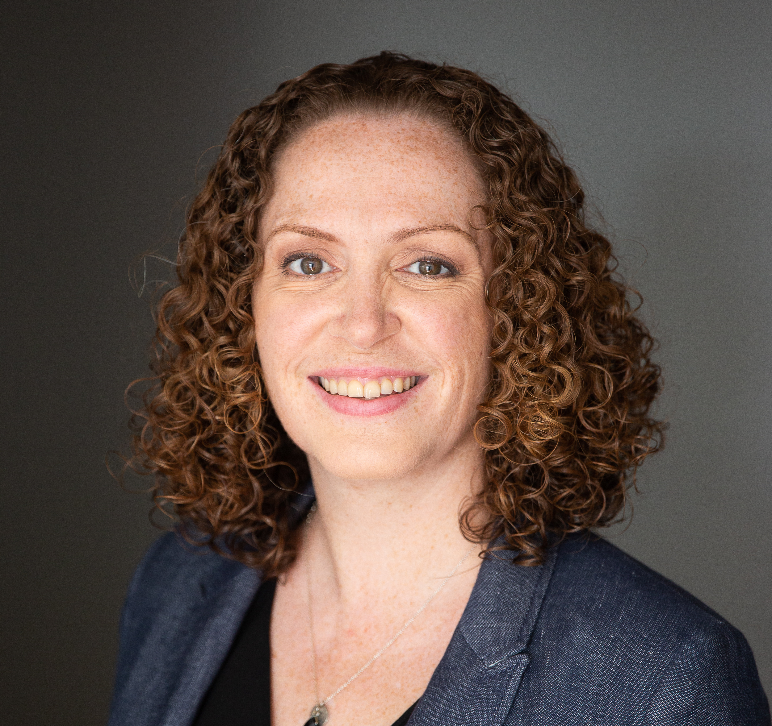 Close-up portrait of a smiling woman with curly red hair, wearing a dark blazer and a black top, against a gray background.