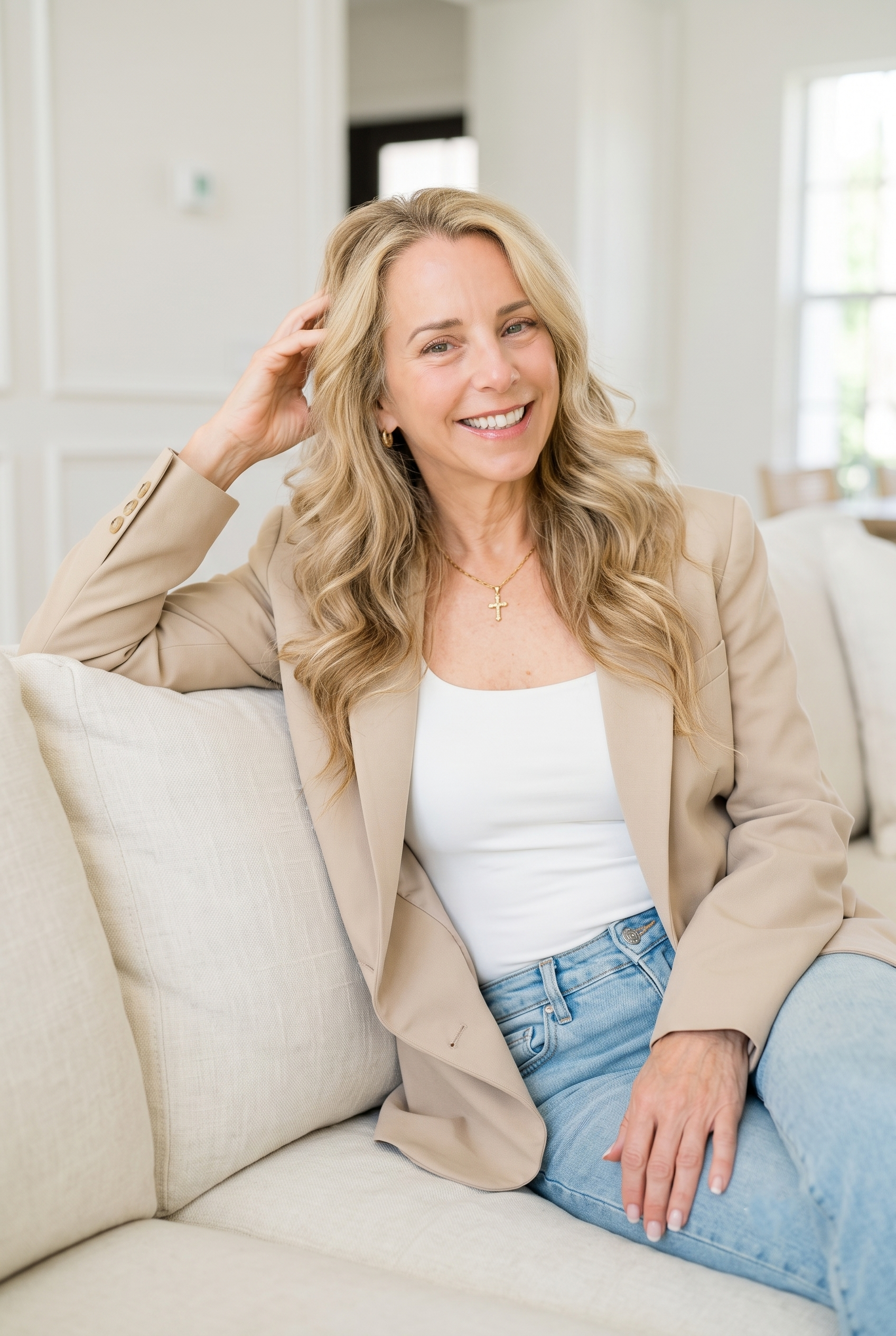 A woman with blonde wavy hair, smiling and wearing a beige blazer over a white top and blue jeans, sitting on a light-colored sofa in a bright room with large windows.