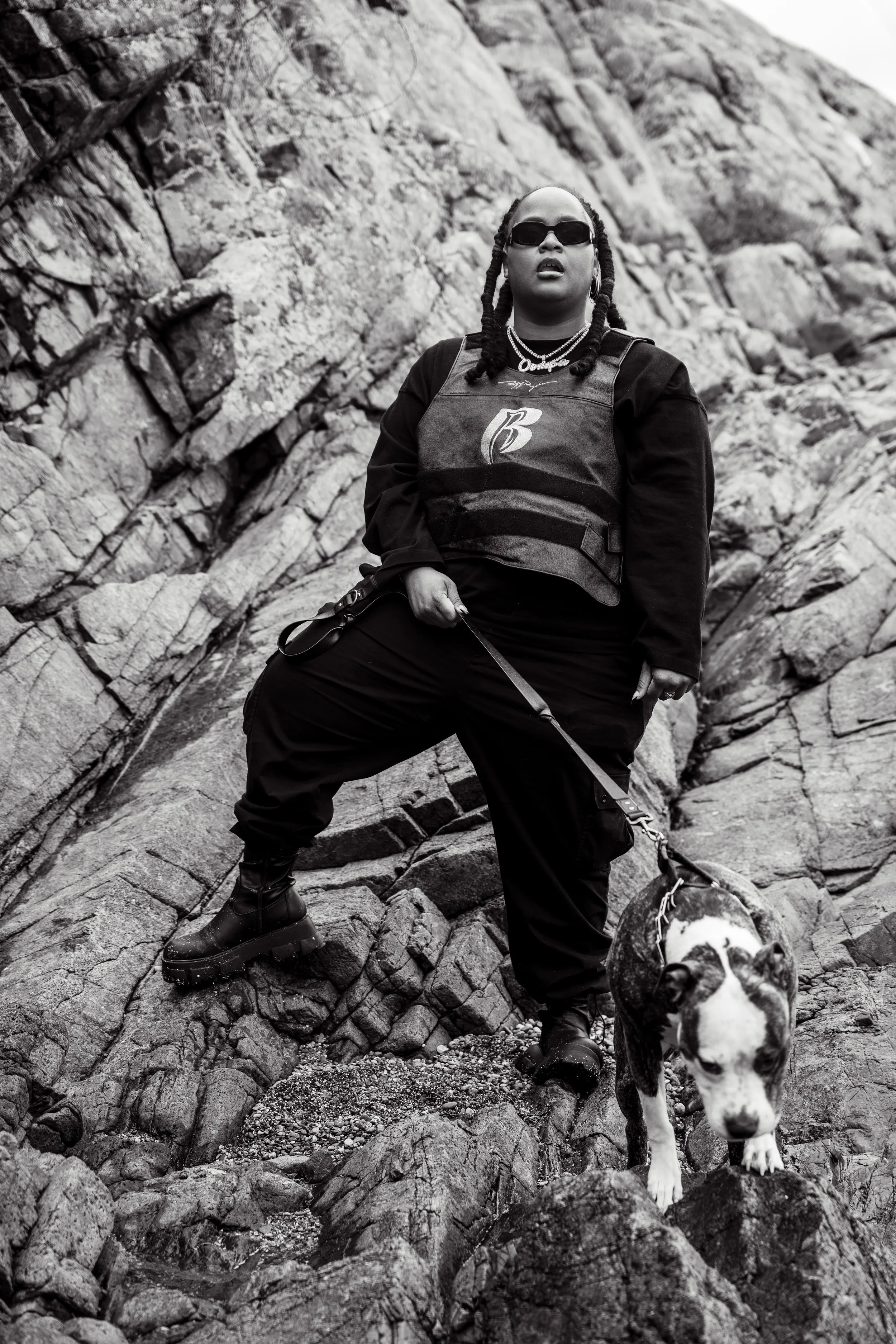 A woman wearing sunglasses, a vest, and black pants, standing on rocky terrain with her dog during a hike.