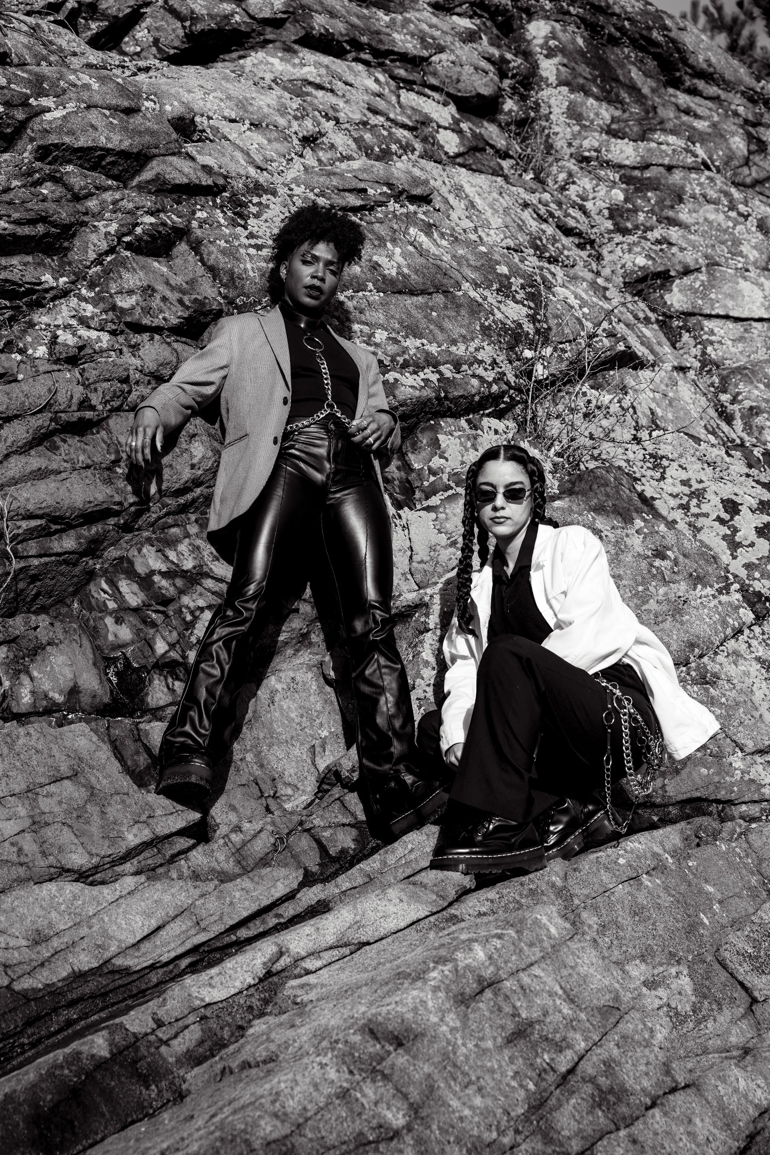 Two women in stylish outfits pose against a rocky outdoor background, one standing and the other crouching, both wearing sunglasses and chain accessories in a black-and-white photo.
