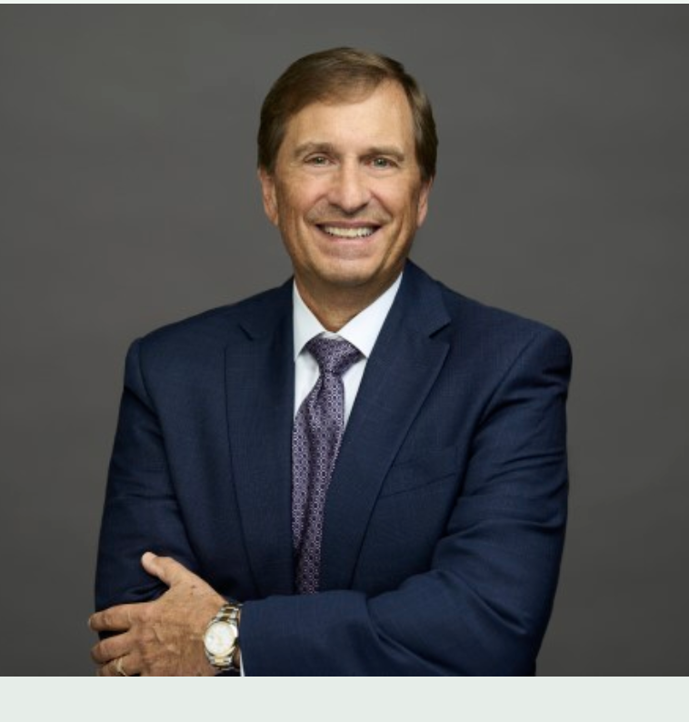 A smiling man in a navy suit with a patterned tie, wearing a watch, standing against a plain gray background.