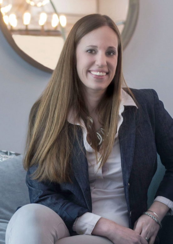 A woman with long light brown hair, smiling, sitting indoors in front of a round mirror with lights, wearing a white shirt and a dark blazer.