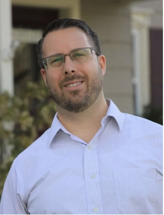 A man with glasses and a beard wearing a light blue collared shirt standing outdoors in front of a house.