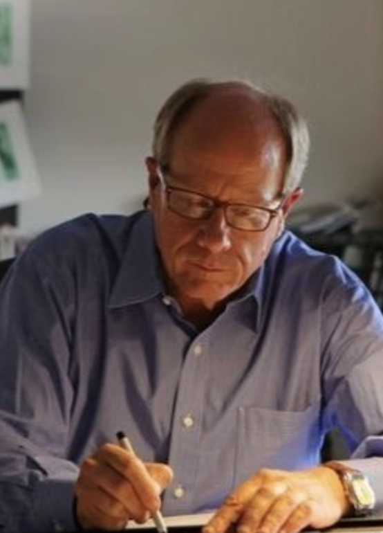 An older man with glasses and a watch, wearing a blue shirt, writing on paper with a pen in an office or study.