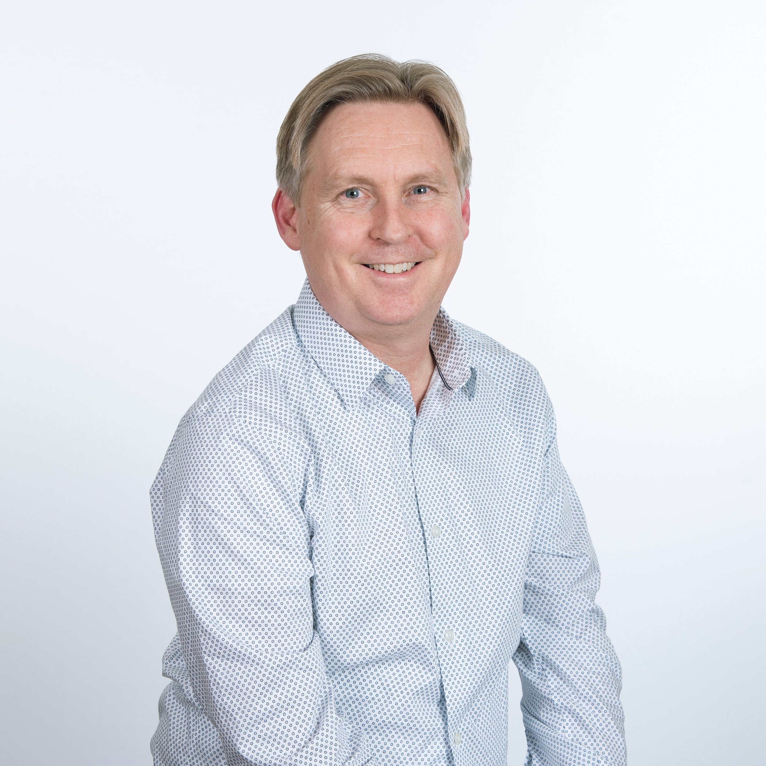 Professional portrait of smiling middle-aged man with blond hair wearing a white patterned button-up shirt against a white background.