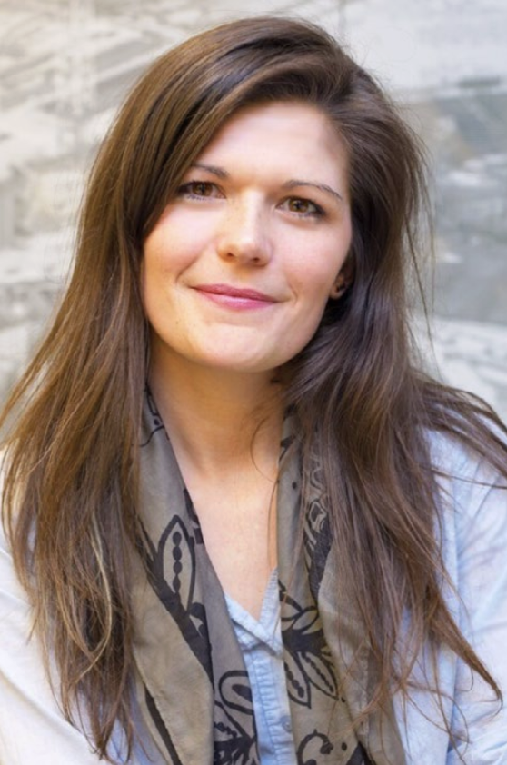 Portrait of a woman with long brown hair wearing a light blue shirt and a patterned scarf, smiling against a stone wall background.