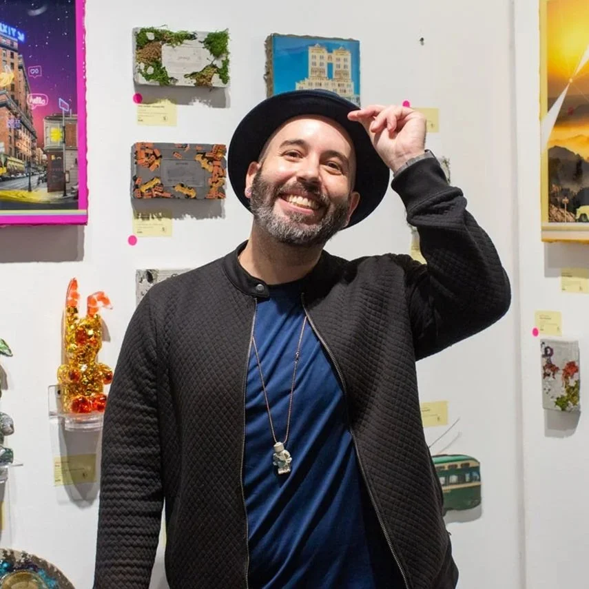 A smiling man with a beard and mustache, wearing a black hat and black quilted jacket, standing in front of a wall with colorful art displays.