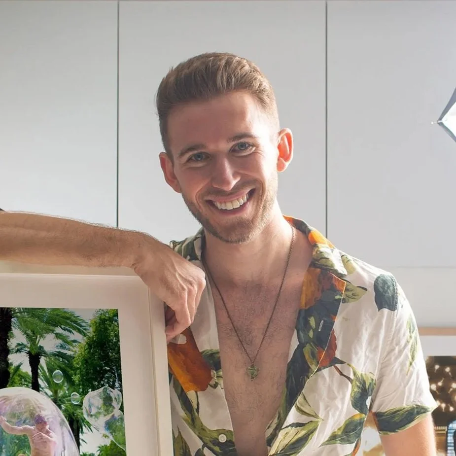 Young man with short hair and a beard, smiling, wearing a tropical print shirt and a necklace, indoors with grey cabinets in the background, holding a framed photo of a person in a lush outdoor setting.