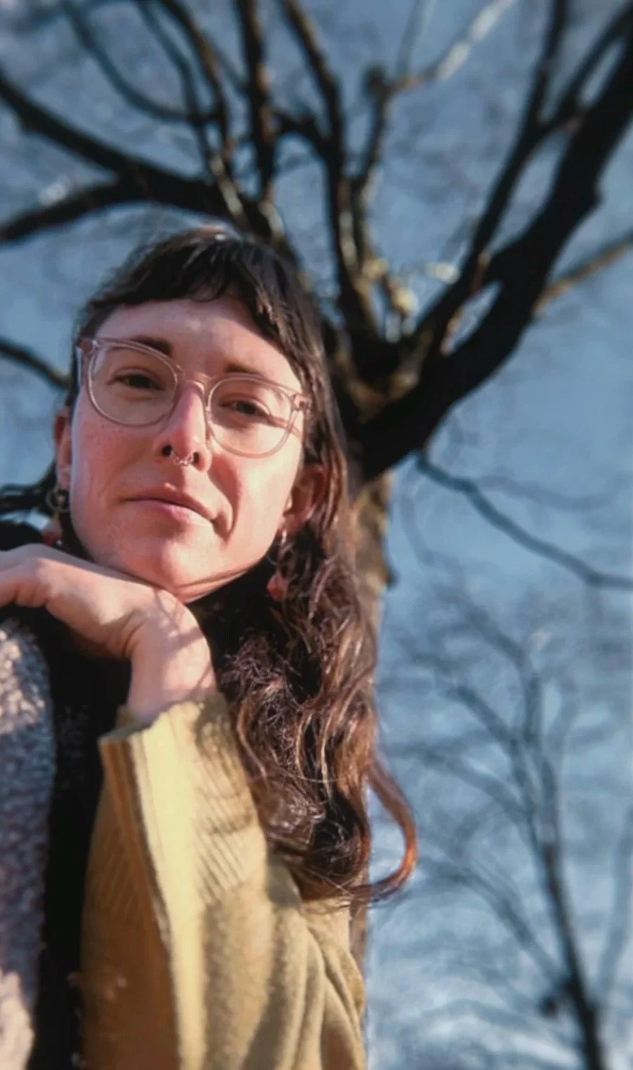 A young woman with glasses and a septum piercing takes a selfie outdoors, with a leafless tree in the background during winter.