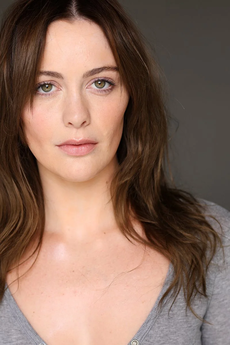 Close-up portrait of a woman with green eyes and long, wavy brown hair, wearing a gray top, against a neutral background.