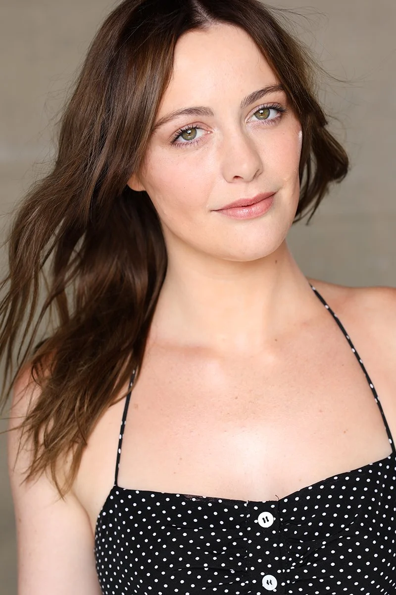 Close-up of a young woman with long, wavy brown hair, light green eyes, and a slight smile, wearing a black spaghetti strap top with white polka dots.
