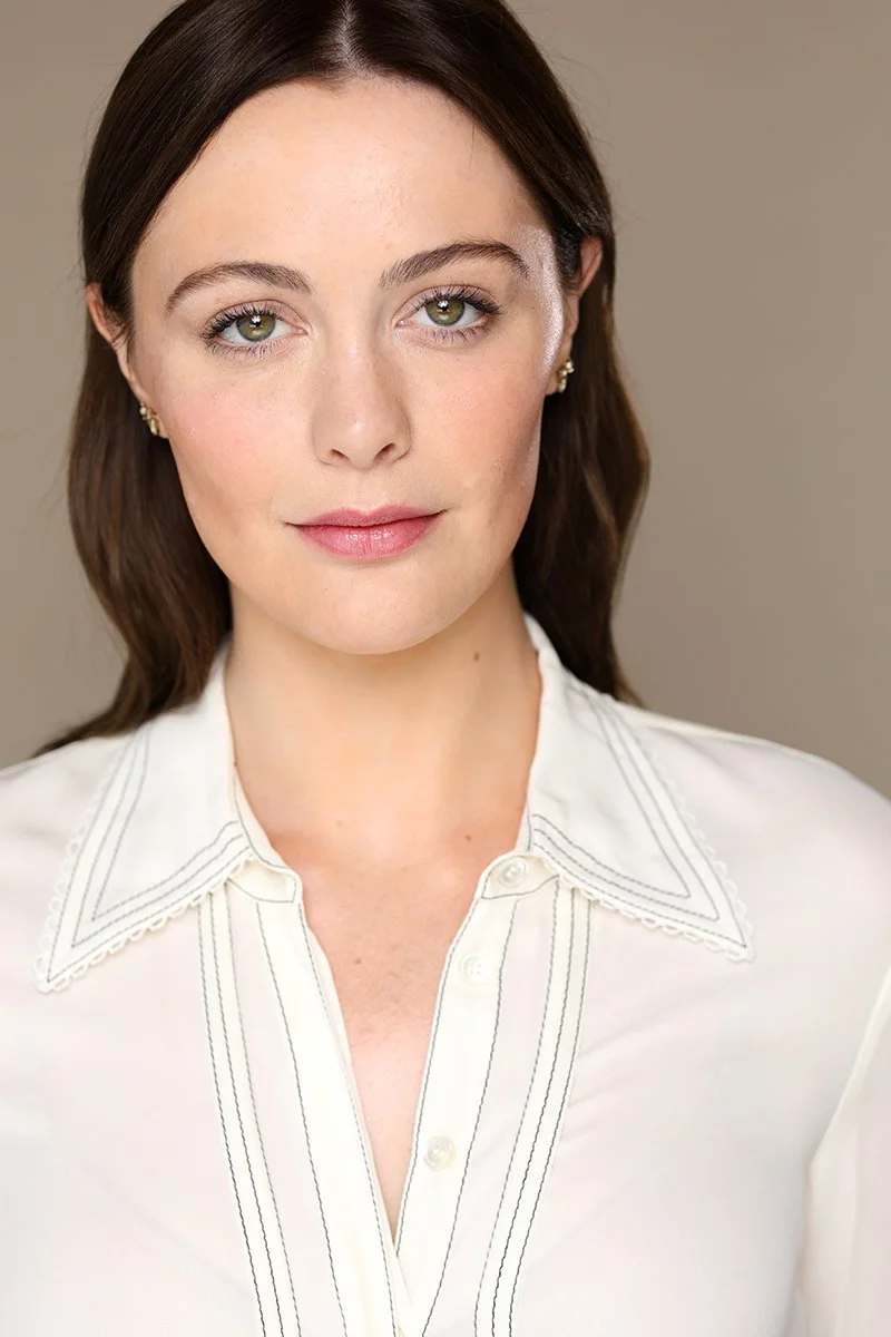 Close-up of a woman with light skin, shoulder-length dark brown hair, green eyes, and natural makeup, wearing a white blouse with embroidered collar and buttons, and small gold earrings, posing against a neutral background.