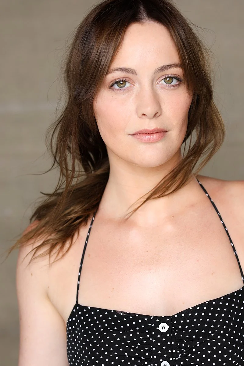 A woman with light skin, green eyes, and brown hair looking directly at the camera with a neutral expression, wearing a black sleeveless dress with white polka dots.