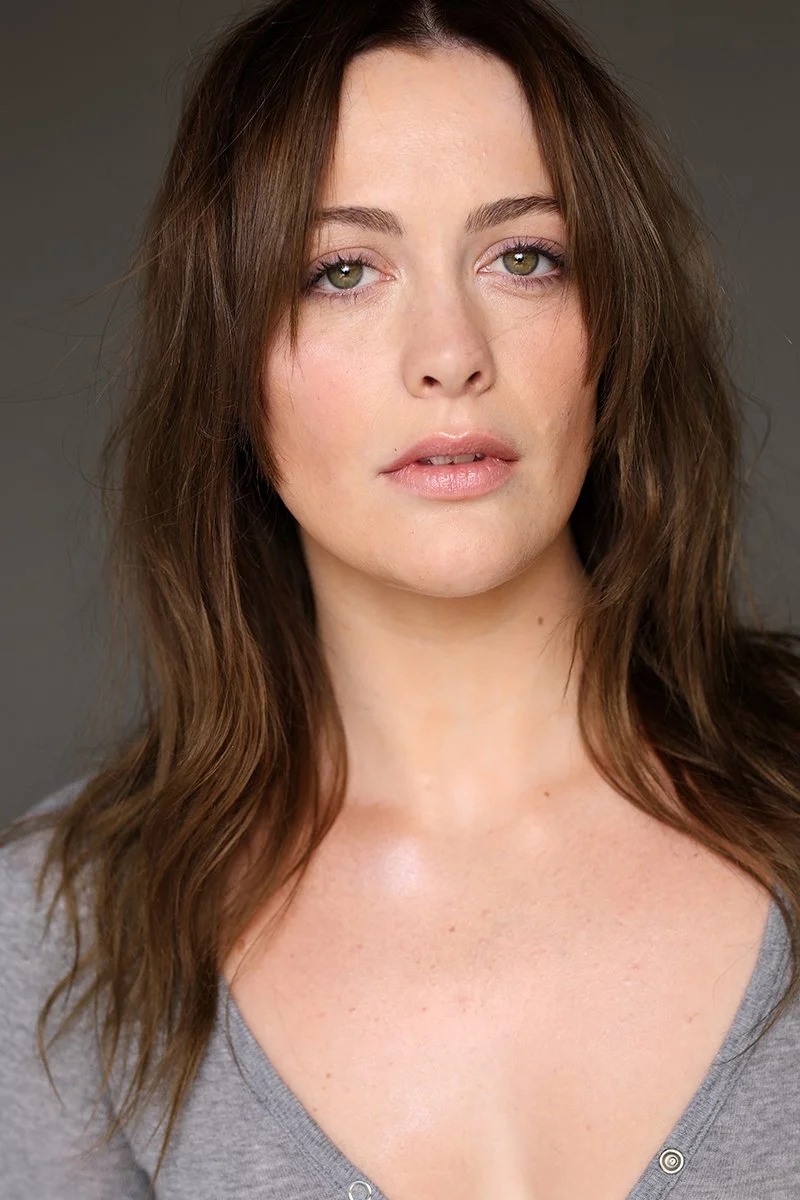 Close-up of a woman with wavy brown hair and green eyes, wearing a gray top, against a neutral background.