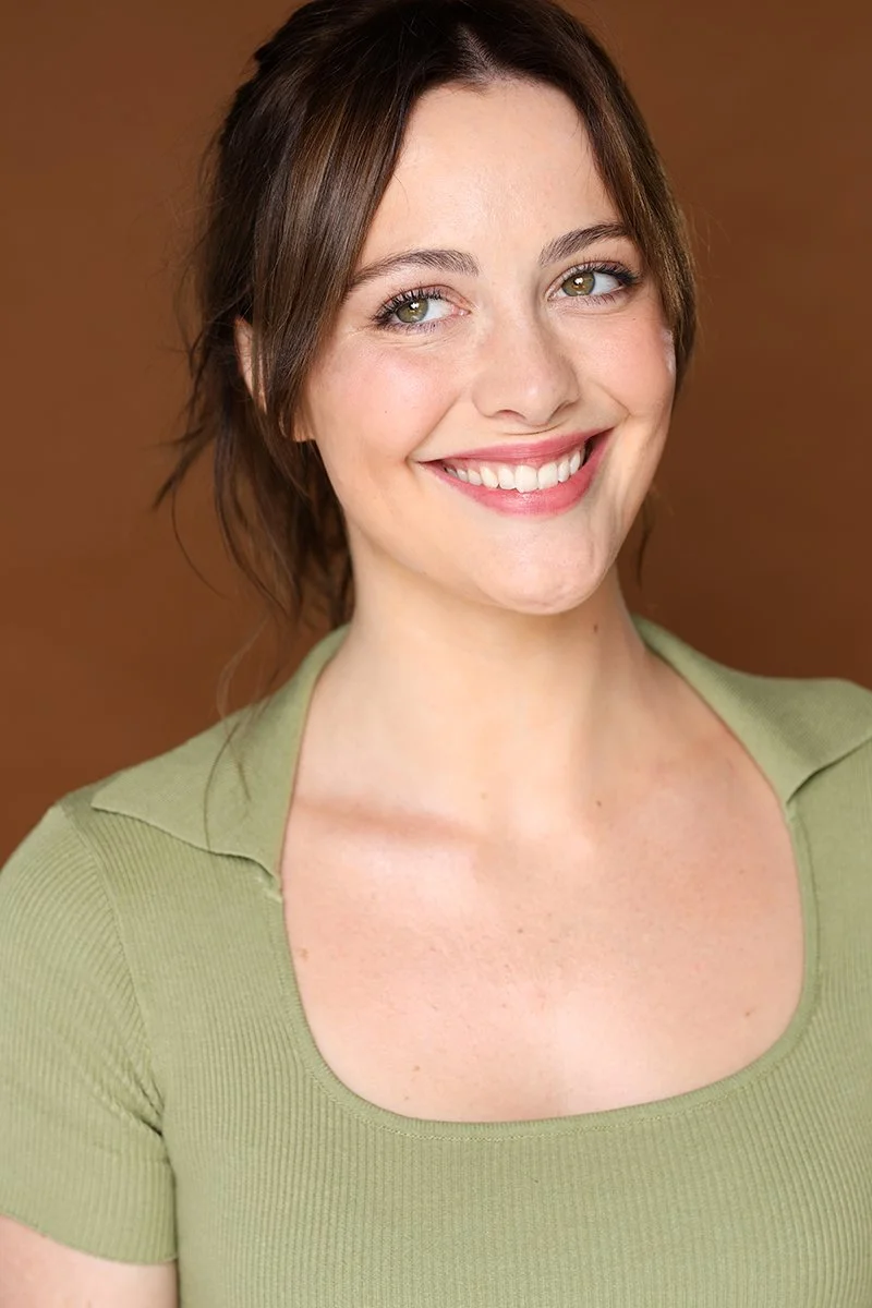 A young woman with light skin, brown hair, and green eyes smiling at the camera, wearing a light green top, against a brown background.