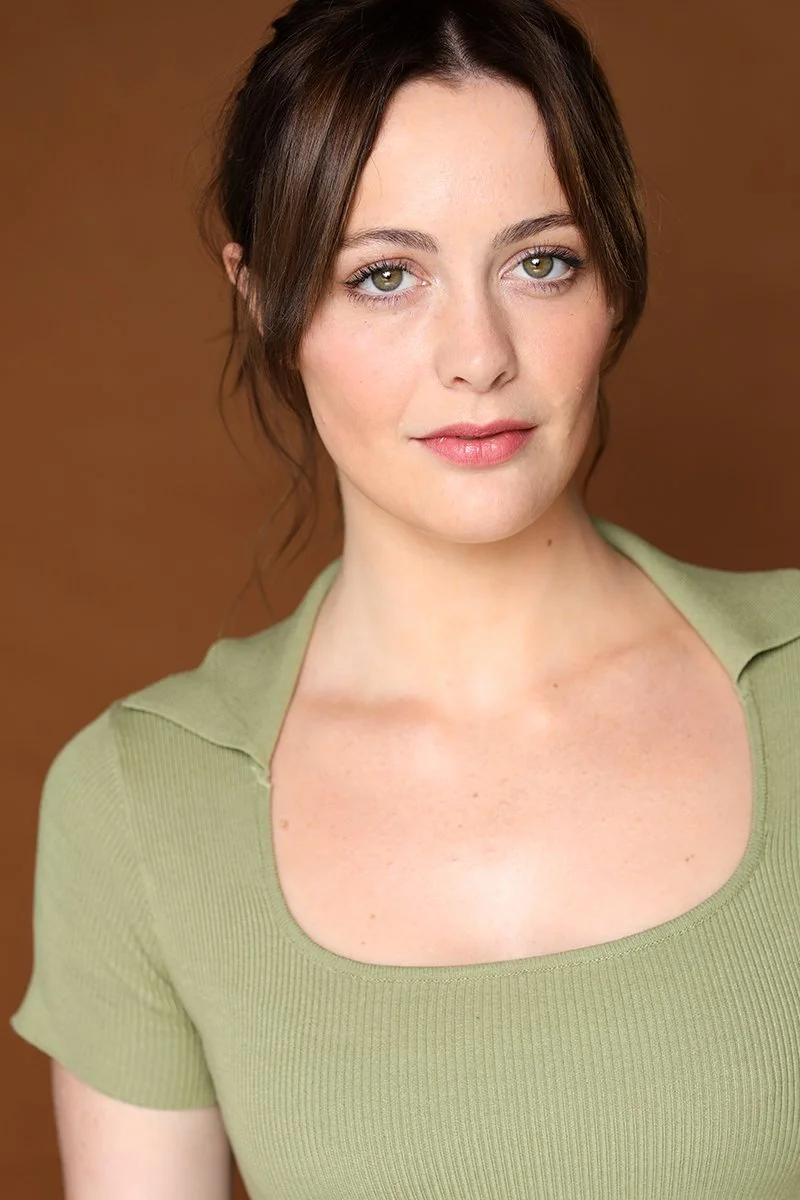 A young woman with fair skin and green eyes, wearing a light green top, looking at the camera against a brown background.