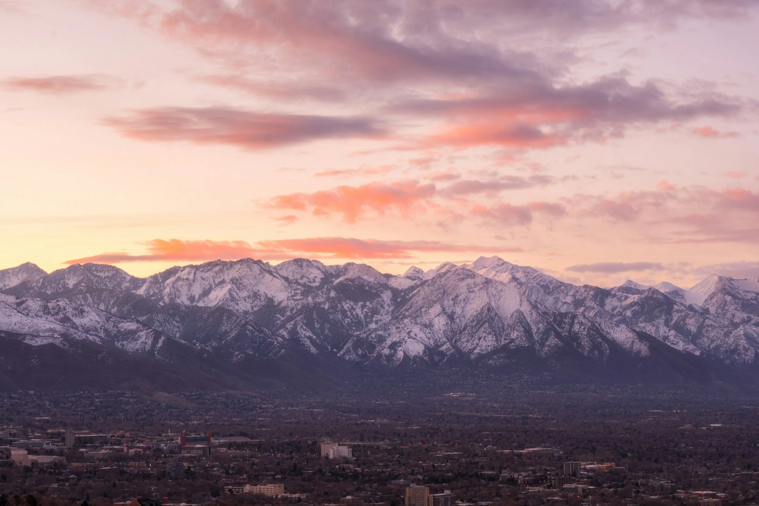 A cityscape with a mountain range in the background during a sunset, with pink and purple clouds in the sky.