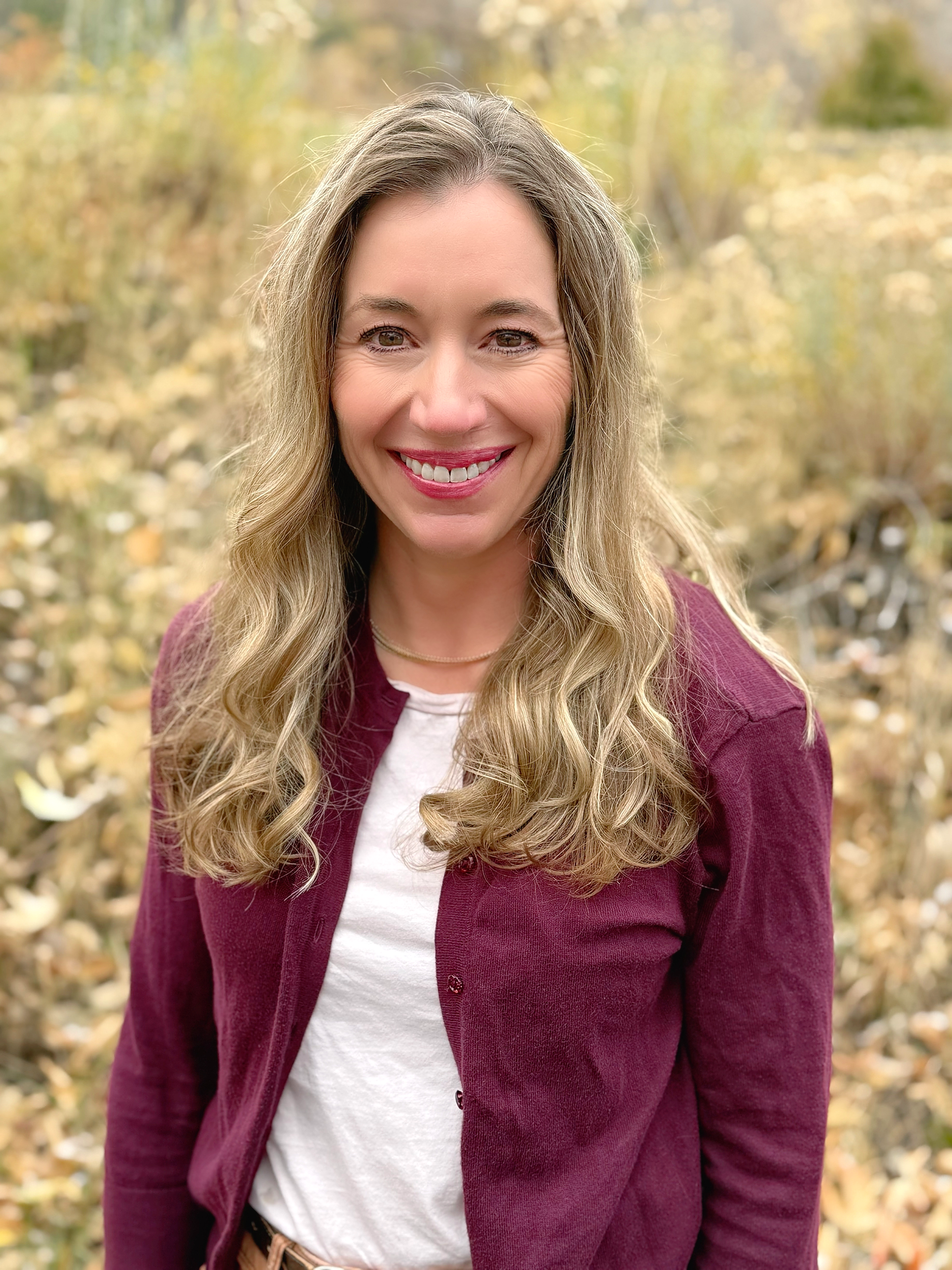 A woman with long, wavy blonde hair smiling outdoors in a fall setting with yellow and orange foliage.