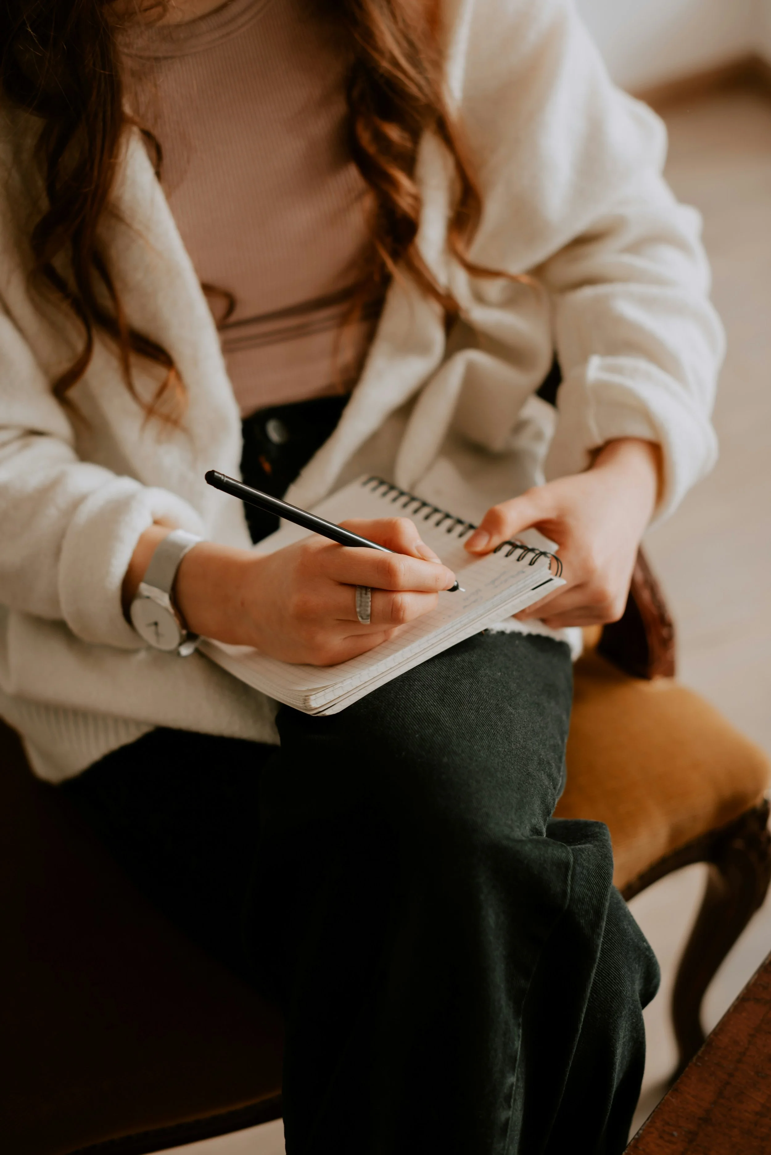 Person sitting on a vintage wooden chair, writing in a spiral notebook with a black pen. They are wearing a beige sweater, a watch, and a ring, with long wavy hair visible.