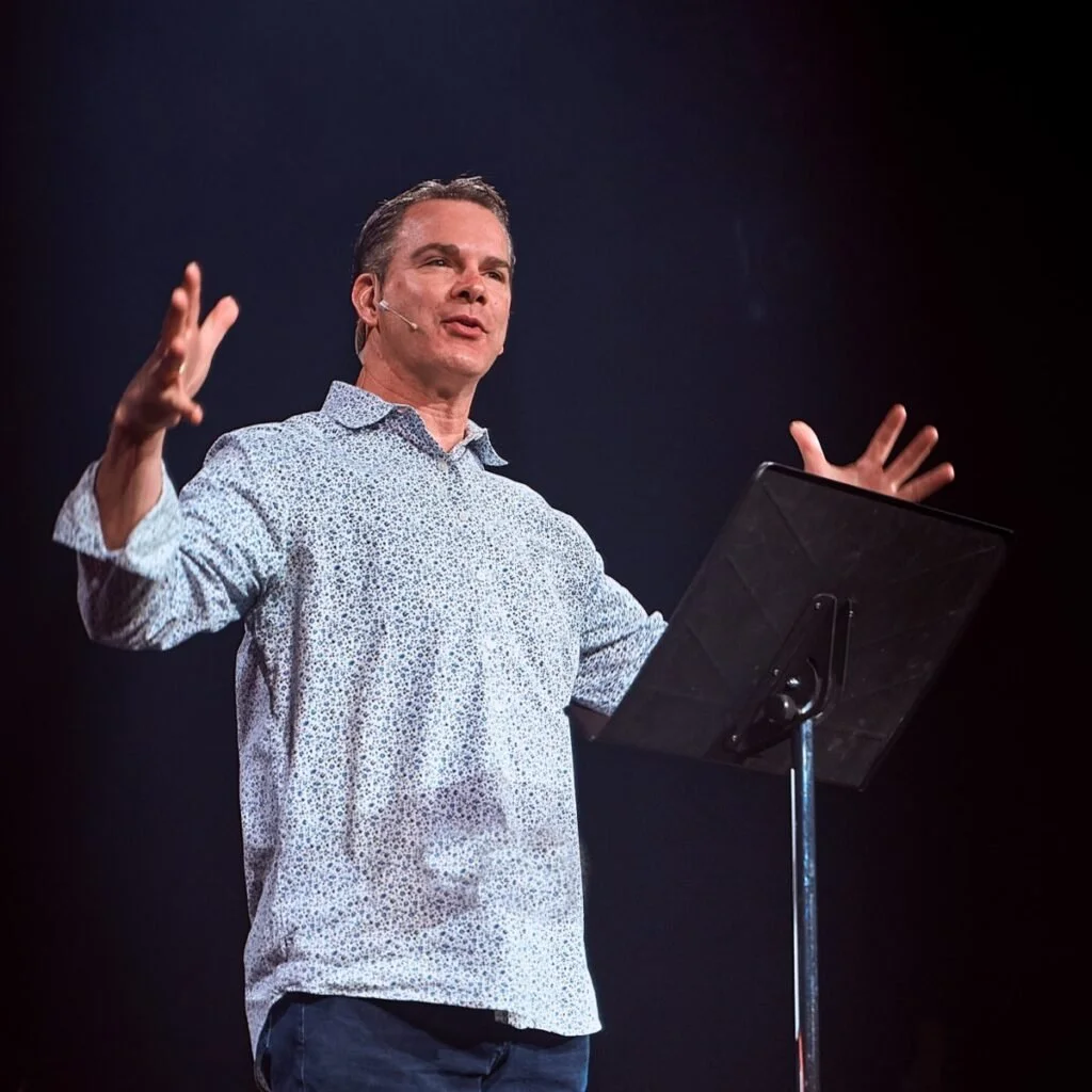 A man giving a speech or presentation on stage with a microphone headset, standing behind a music stand or lectern.