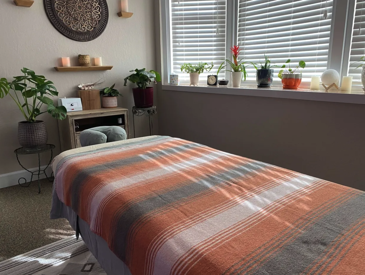 Cozy bedroom with a bed covered in an orange, gray, and cream plaid blanket, near a large window with white blinds. The window sill holds various potted plants, candles, and small decorative items. A beige wall with a circular wall hanging and floati