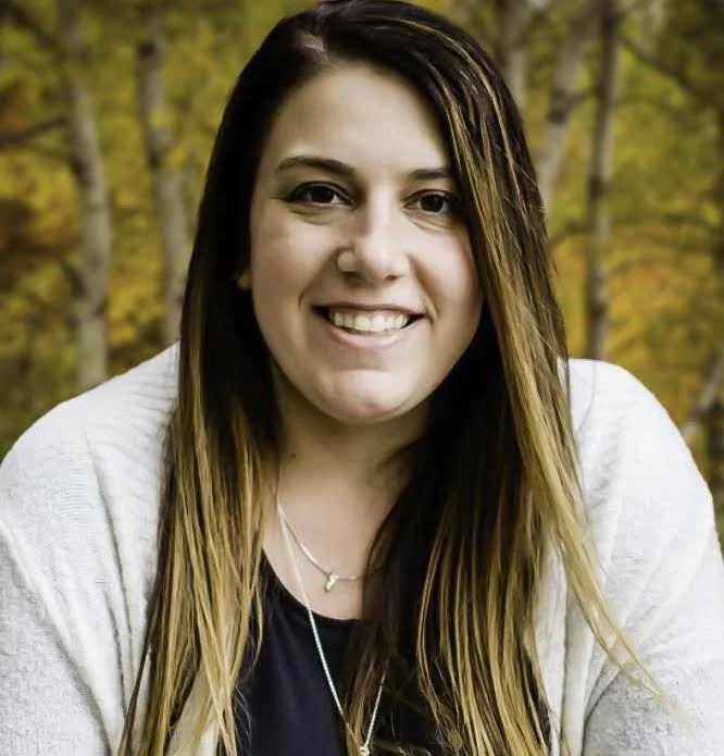 A woman with long, dark brown hair smiling outdoors during fall with yellow and orange leaves in the background.