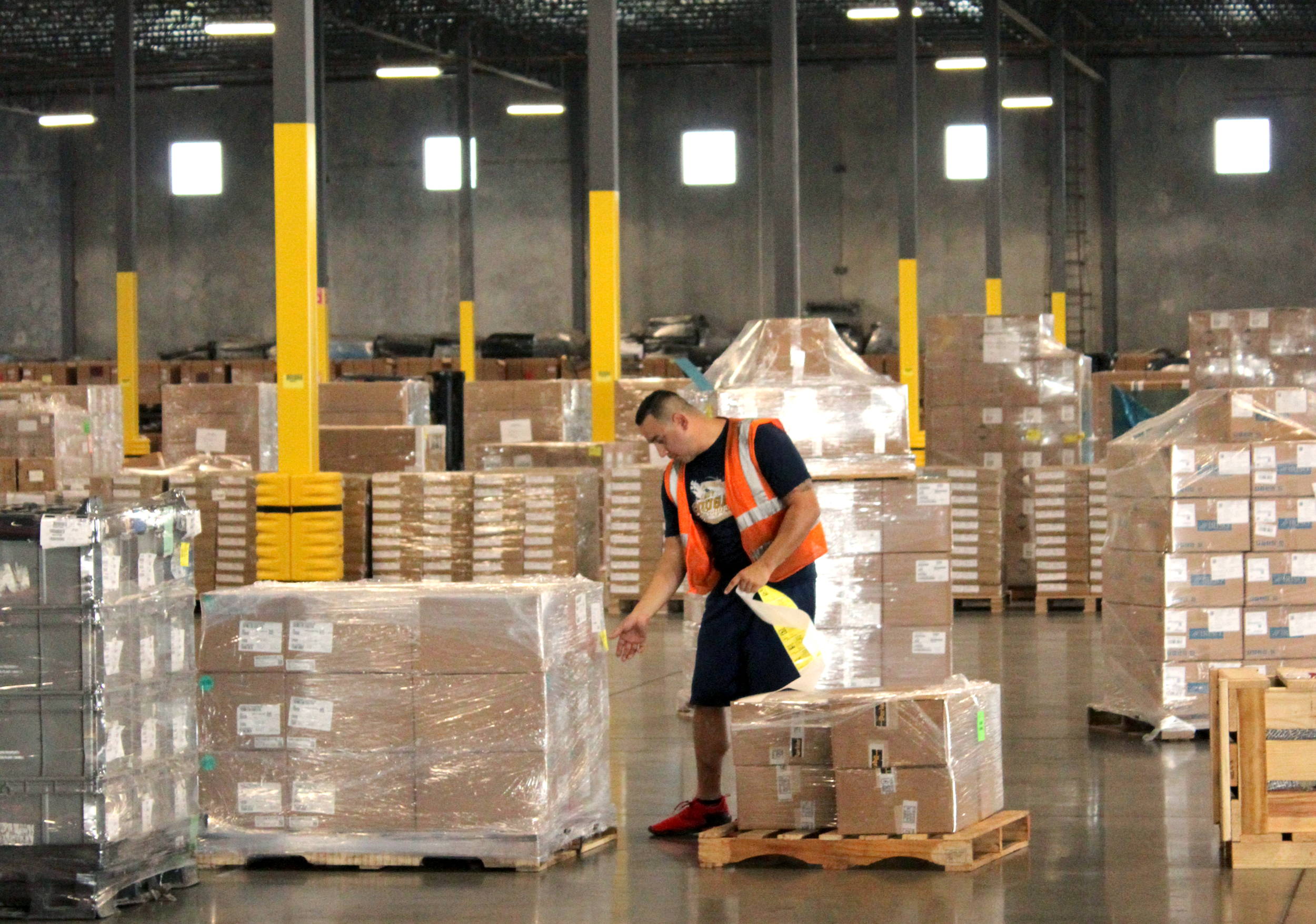 A worker in a warehouse wearing a safety vest and shorts, inspecting pallets of boxed goods wrapped in plastic, with more pallets and warehouse structure in the background.