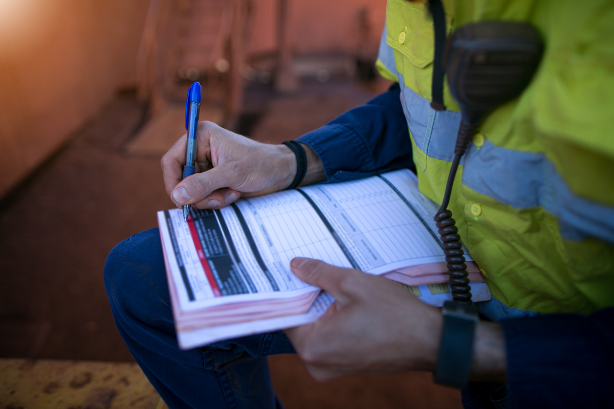 A person wearing a yellow safety vest and navy blue shirt sitting outdoors, holding a logbook and writing with a blue pen. The logbook has multiple columns and text, indicating it might be used for recording data, likely in a work or inspection setting.