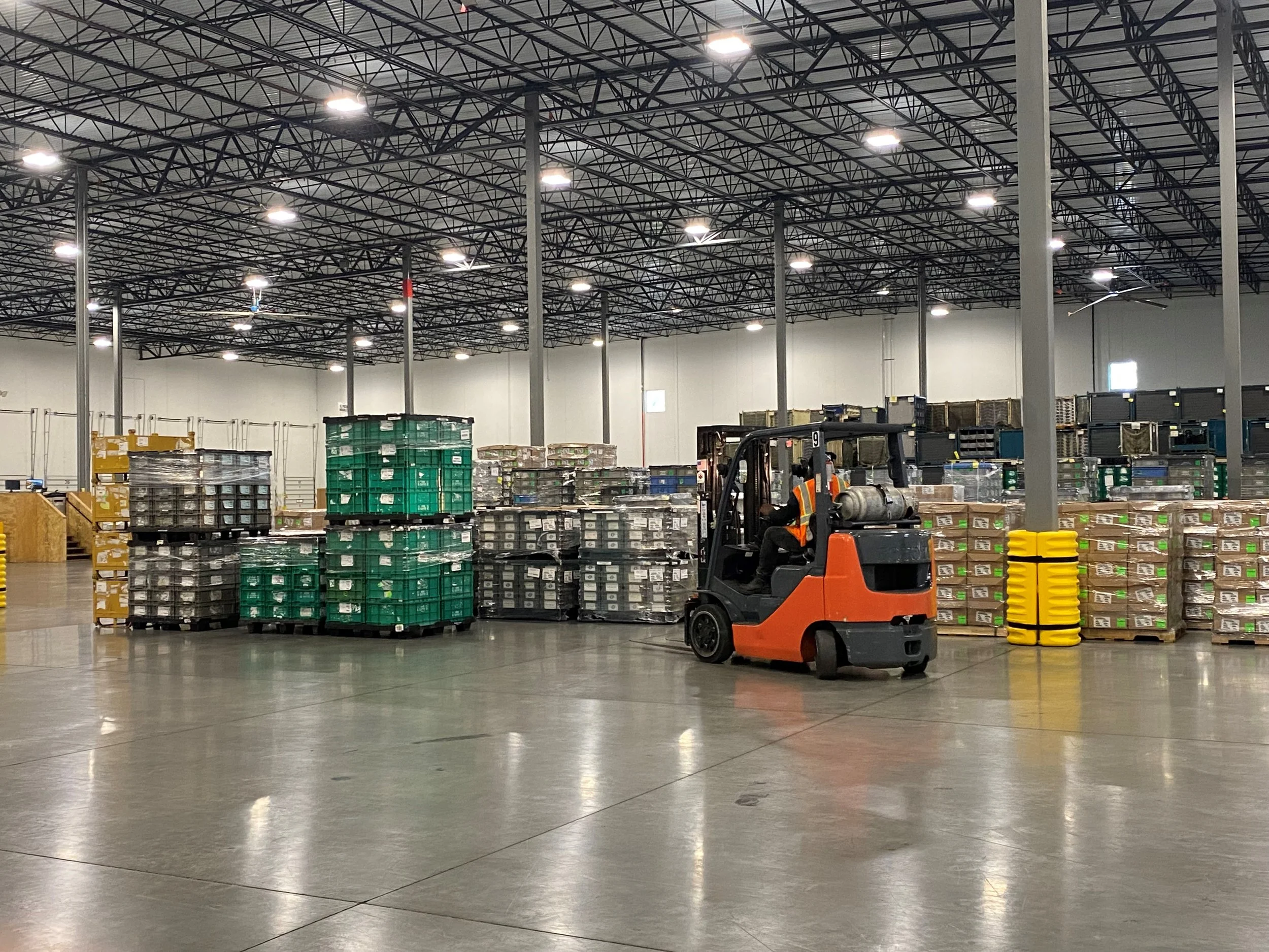 Industrial warehouse with high ceiling, filled with pallets of goods, a forklift operating, and bright overhead lighting.