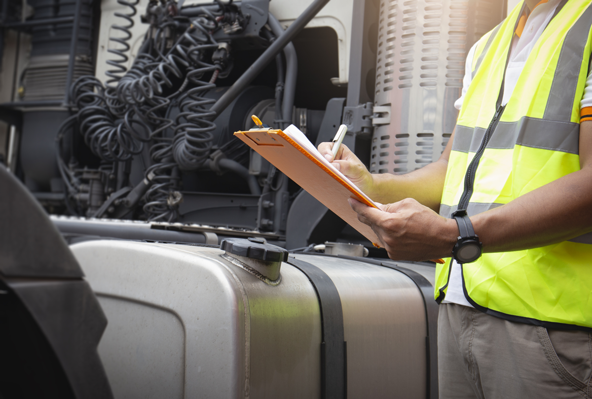 A person wearing a yellow safety vest holding a clipboard and pen, inspecting a large truck engine.