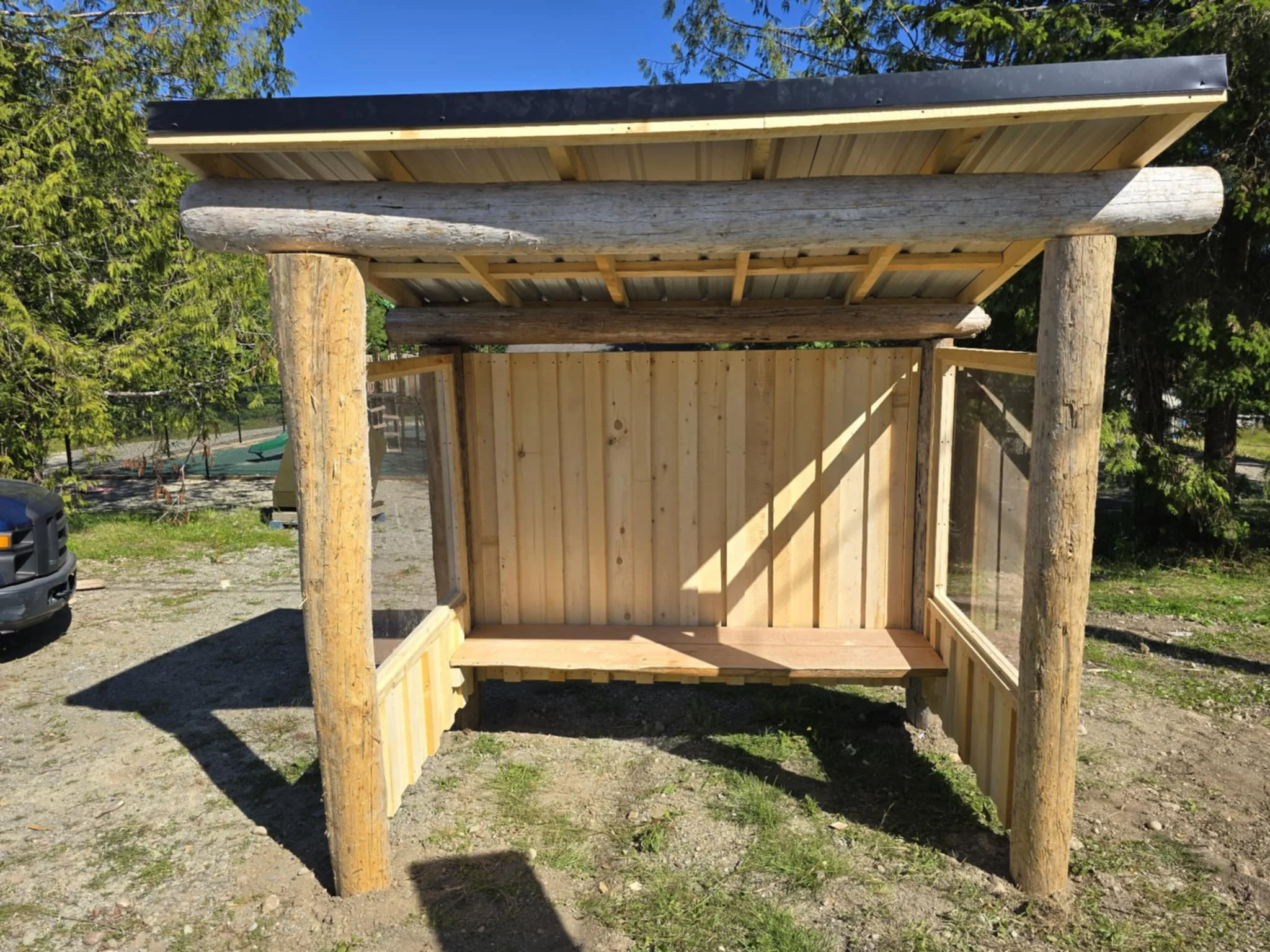 A small wooden outdoor shelter with a slanted roof, supported by four thick wooden logs, and enclosed with wooden and transparent panel walls. It has a bench on one side and is situated on a dirt-ground area with trees and a playground in the background.