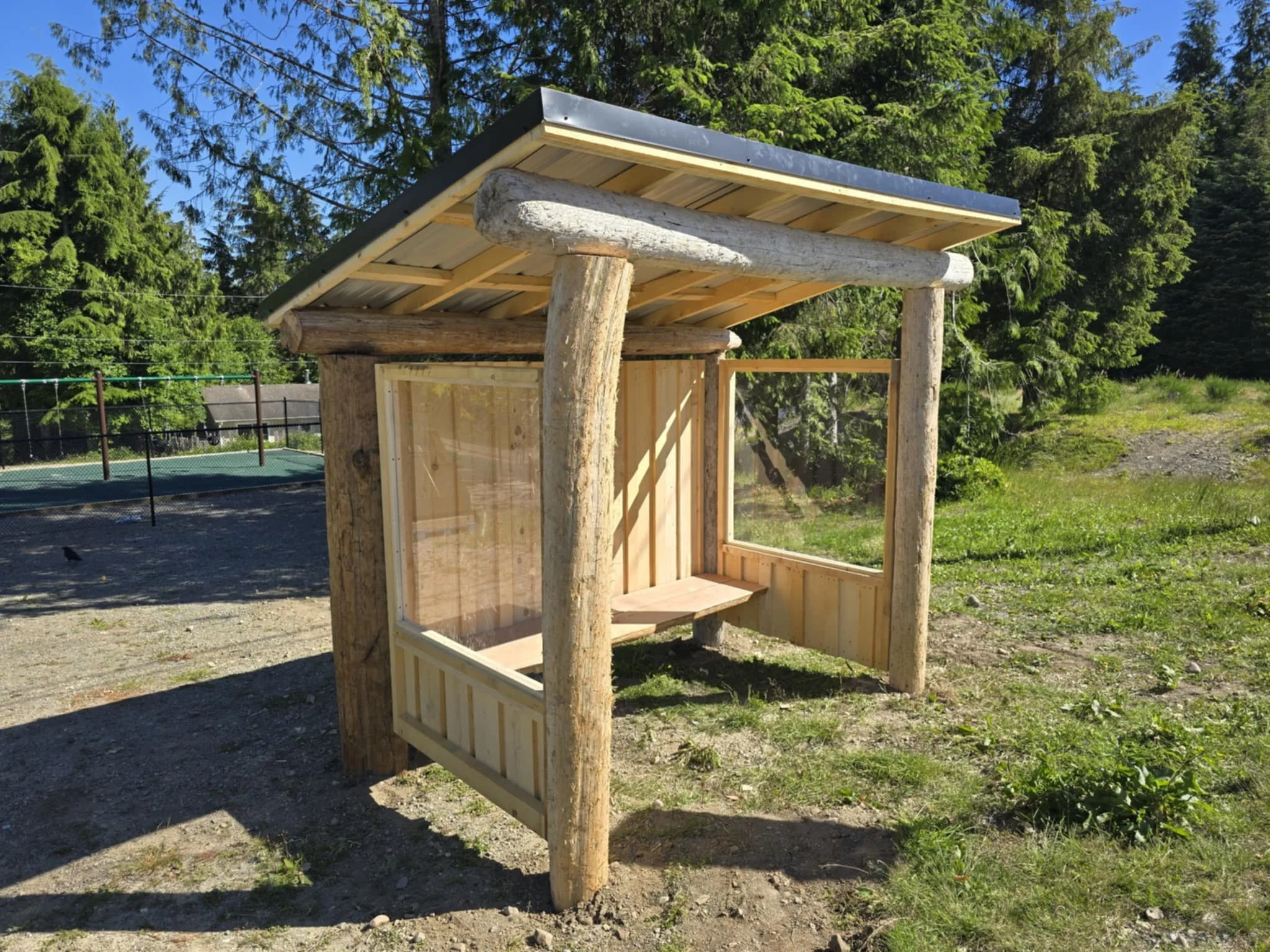 A small wooden shelter with a slanted metal roof and open sides, situated outdoors among trees and grass.