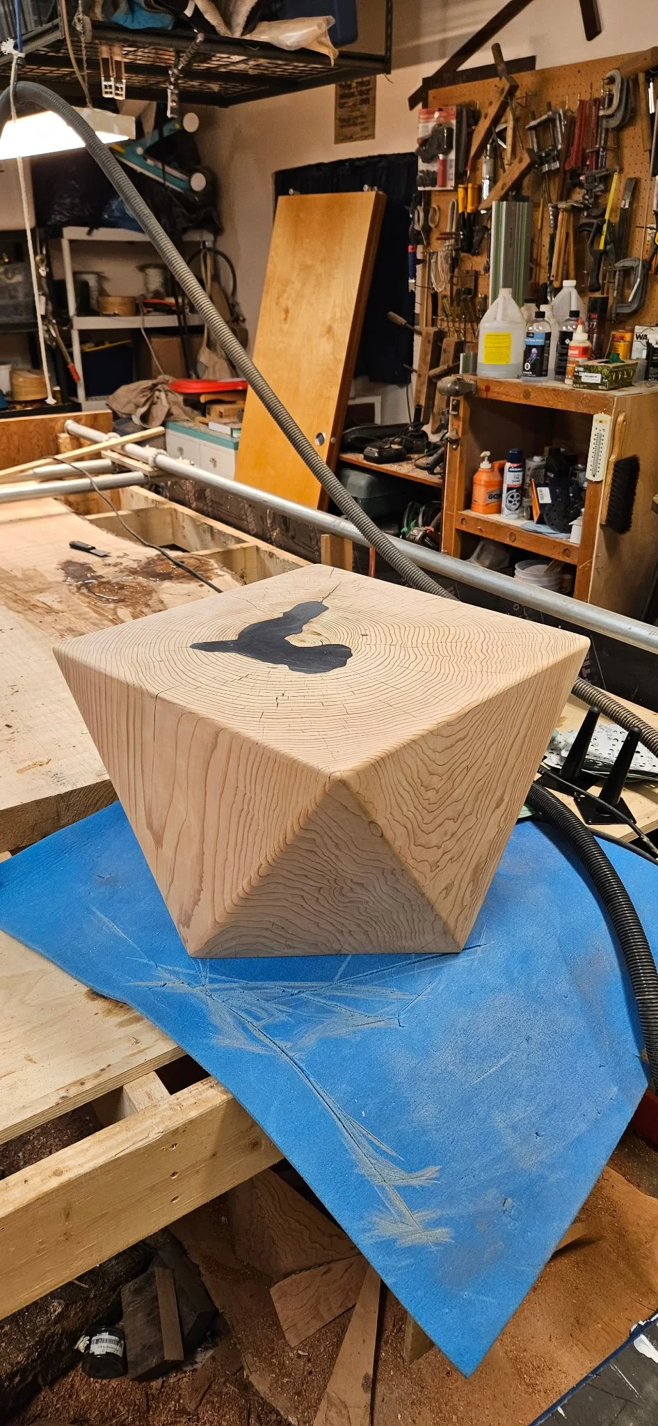 A wooden cube with a black silhouette of a bird painted on the top, sitting on a blue cloth on a workbench in a woodworking shop.