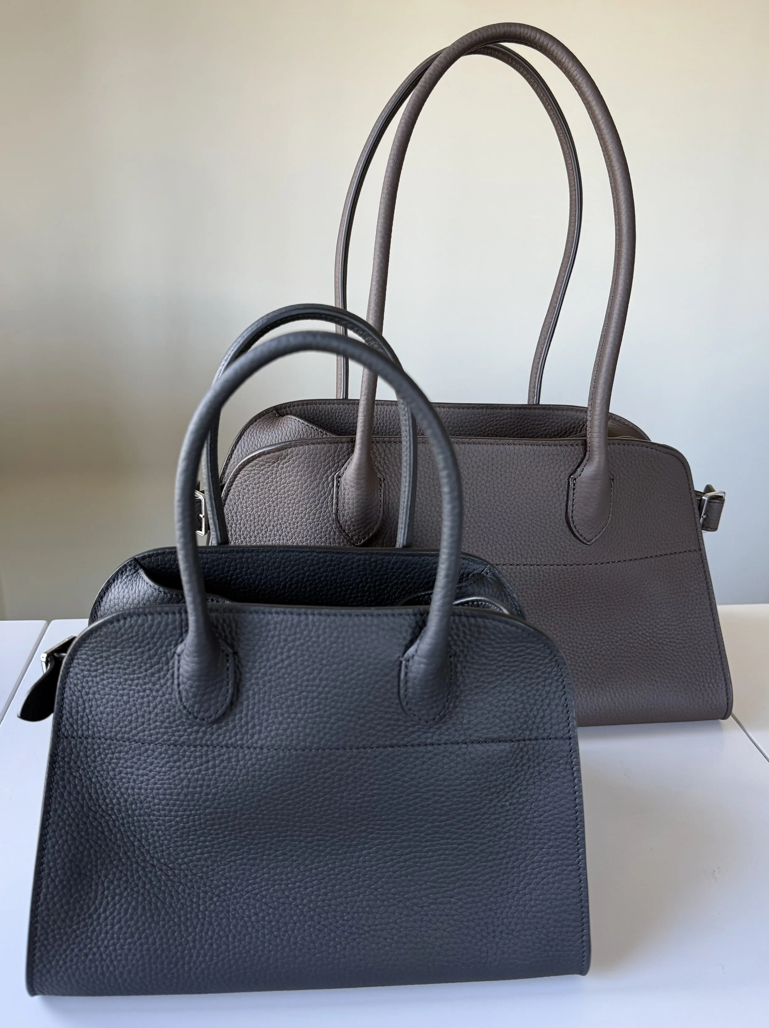 Front view of The Row Margaux 10 in black and Margaux Shoulder 12 in deep brown displayed on a table — showing relative size and structure.