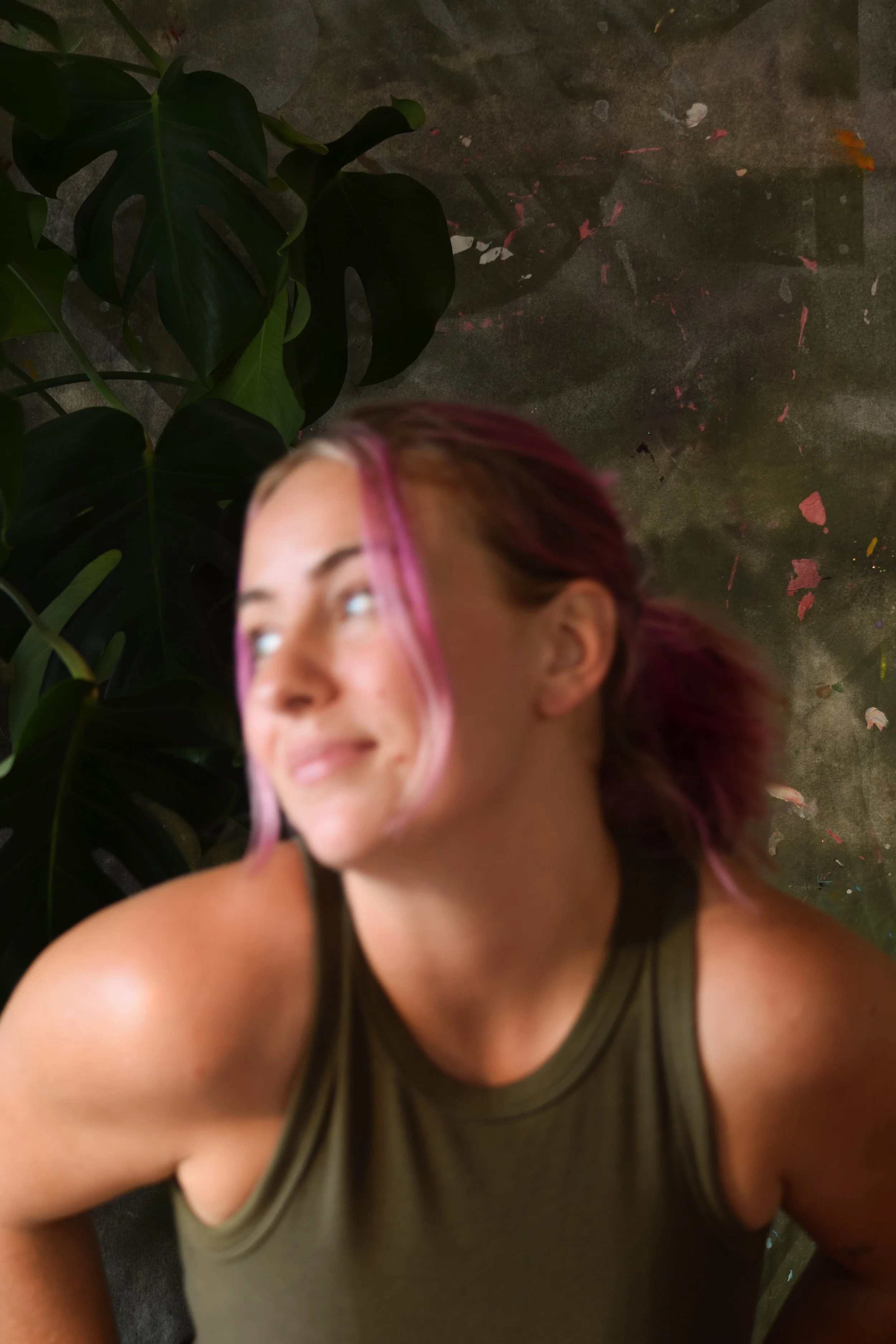 A woman with pink and brown hair smiling and looking to the side, wearing a sleeveless green top, standing near large green leaves against a dark, textured background with paint splatters.