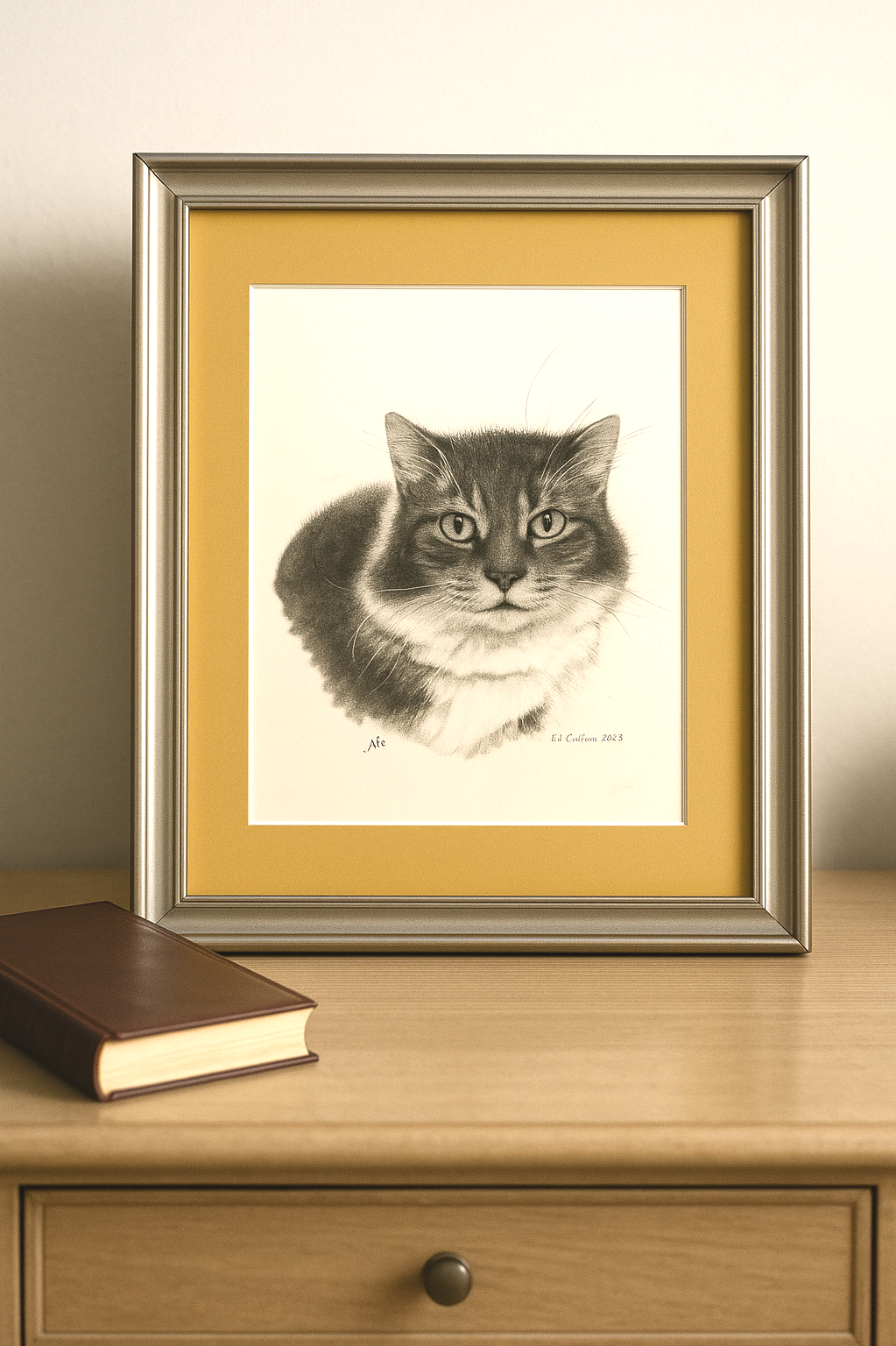 Framed black-and-white pencil drawing of a cat on a wooden surface with a closed brown book nearby.